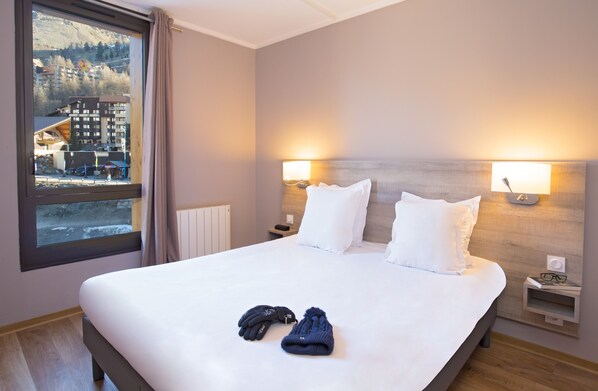 A hotel room with a large bed, two bedside lamps, a view of a mountainous area, and a window with curtains. - SOWELL RÉSIDENCES New Chastillon (Isola)