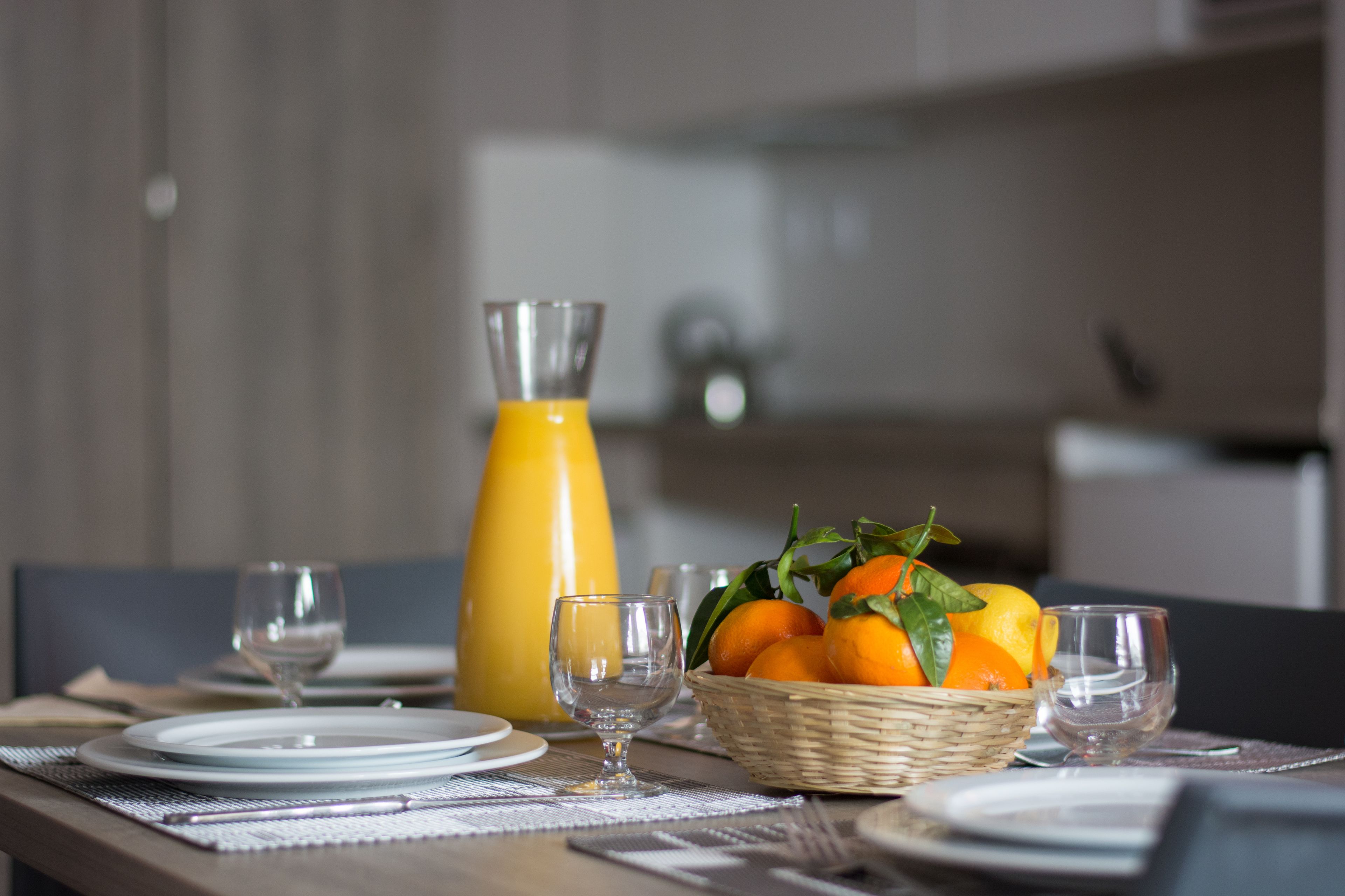 A dining table set with plates, glasses, a glass pitcher, and a basket of oranges.