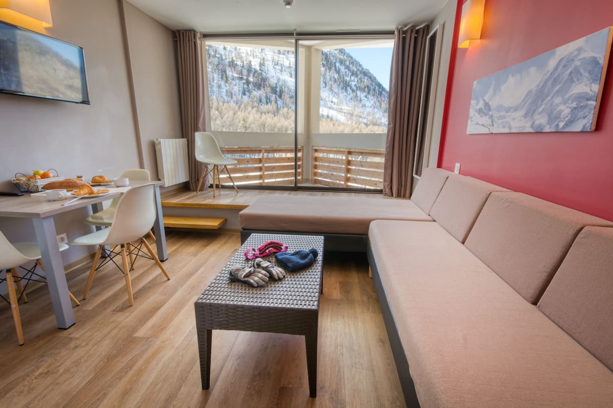 Une chambre d’hôtel moderne avec un plancher en bois, un canapé, une table à manger avec des chaises, une petite table sur laquelle sont posées une paire de chaussons, et une grande fenêtre donnant sur la montagne.
