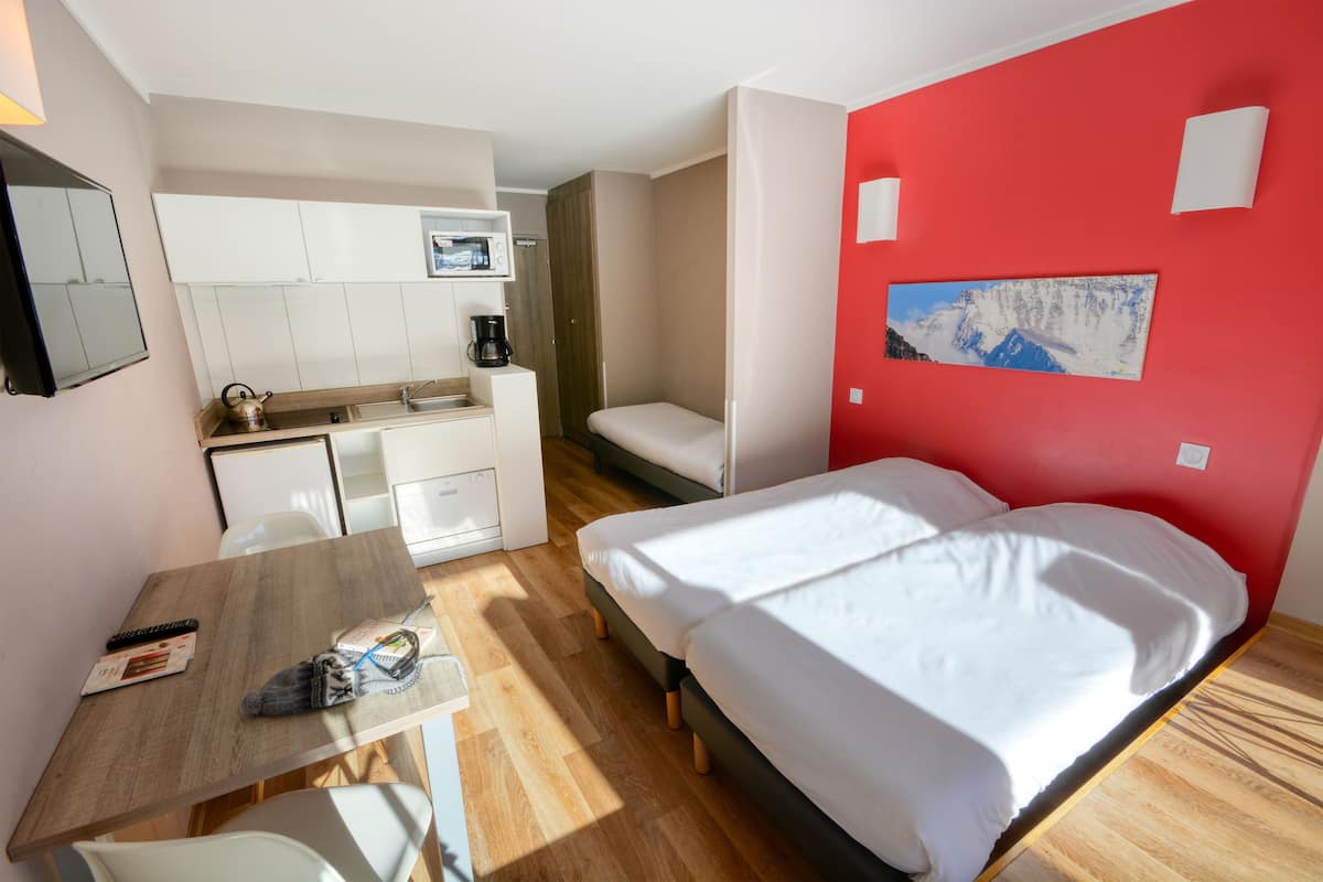 Une pièce compacte comprenant un lit, un petit coin cuisine, une table à manger et un mur d’accent rouge.