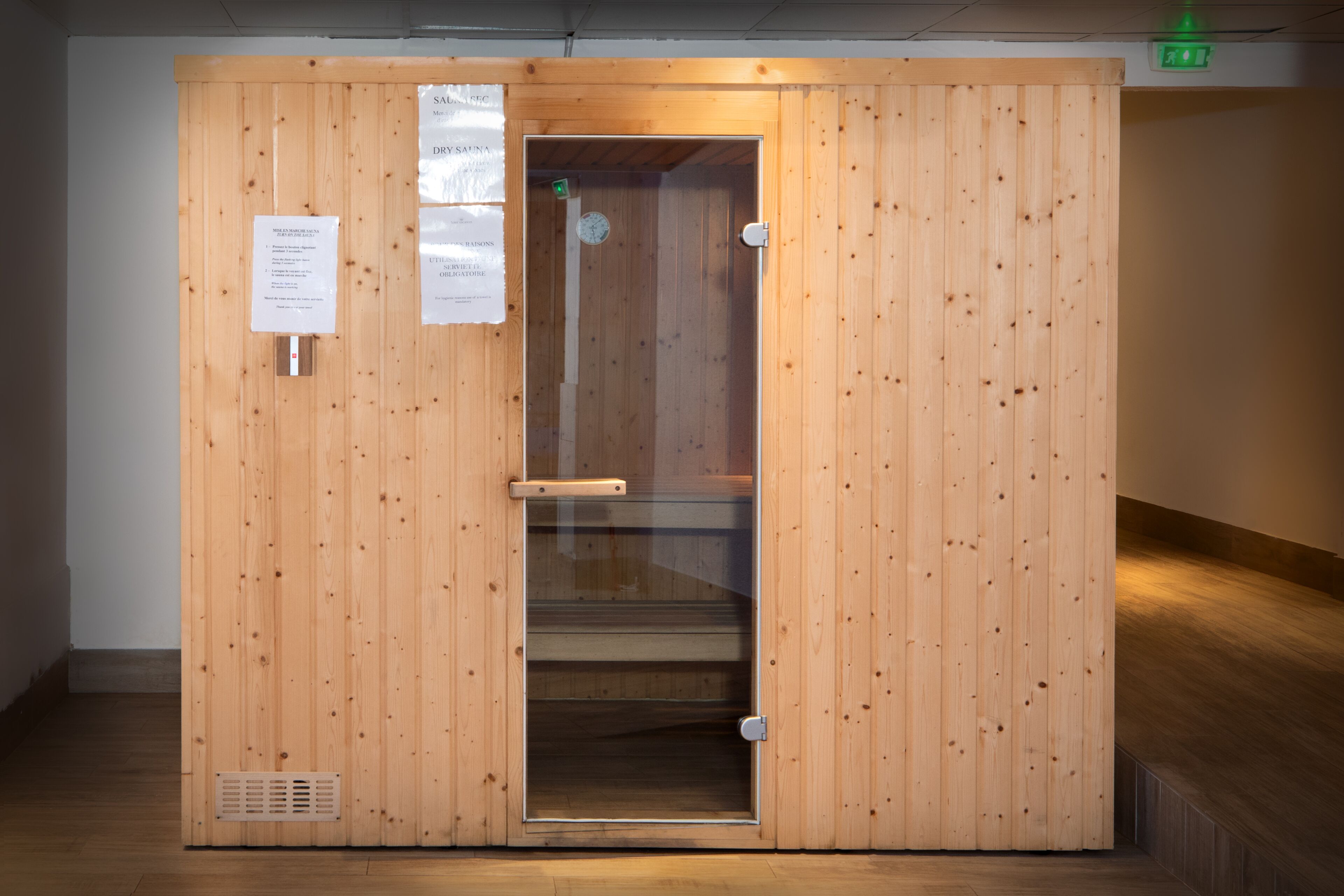 A wooden sauna room with a glass door, ventilation system, and informational signs.
