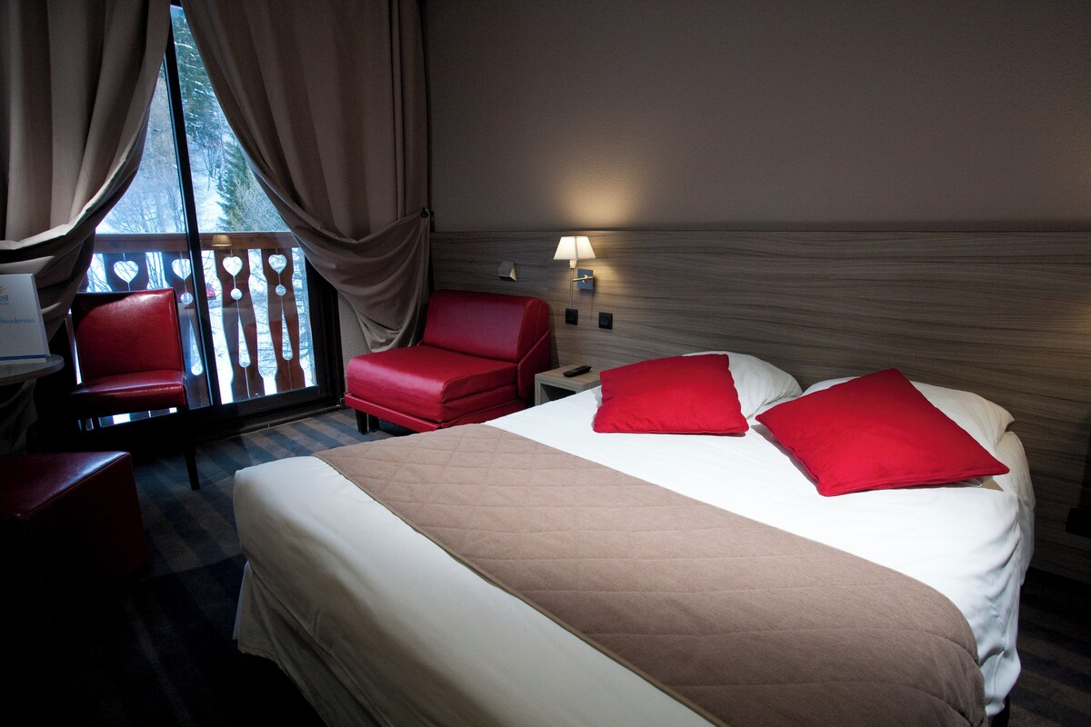 A hotel room with a large bed, red chairs, a desk, and a window with curtains.
