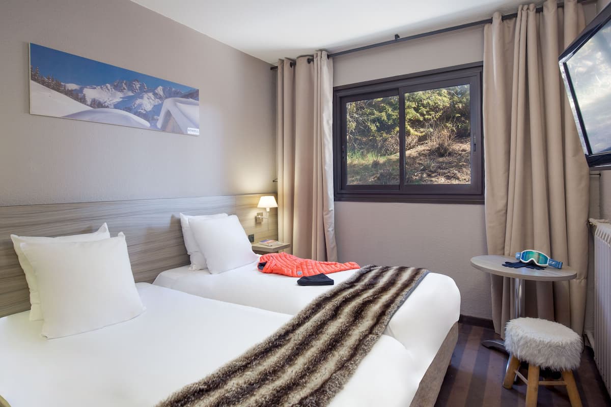 Une chambre d’hôtel avec deux lits, une fenêtre avec des rideaux, une petite table sur laquelle se trouve une paire de lunettes et une lampe sur la table de chevet.