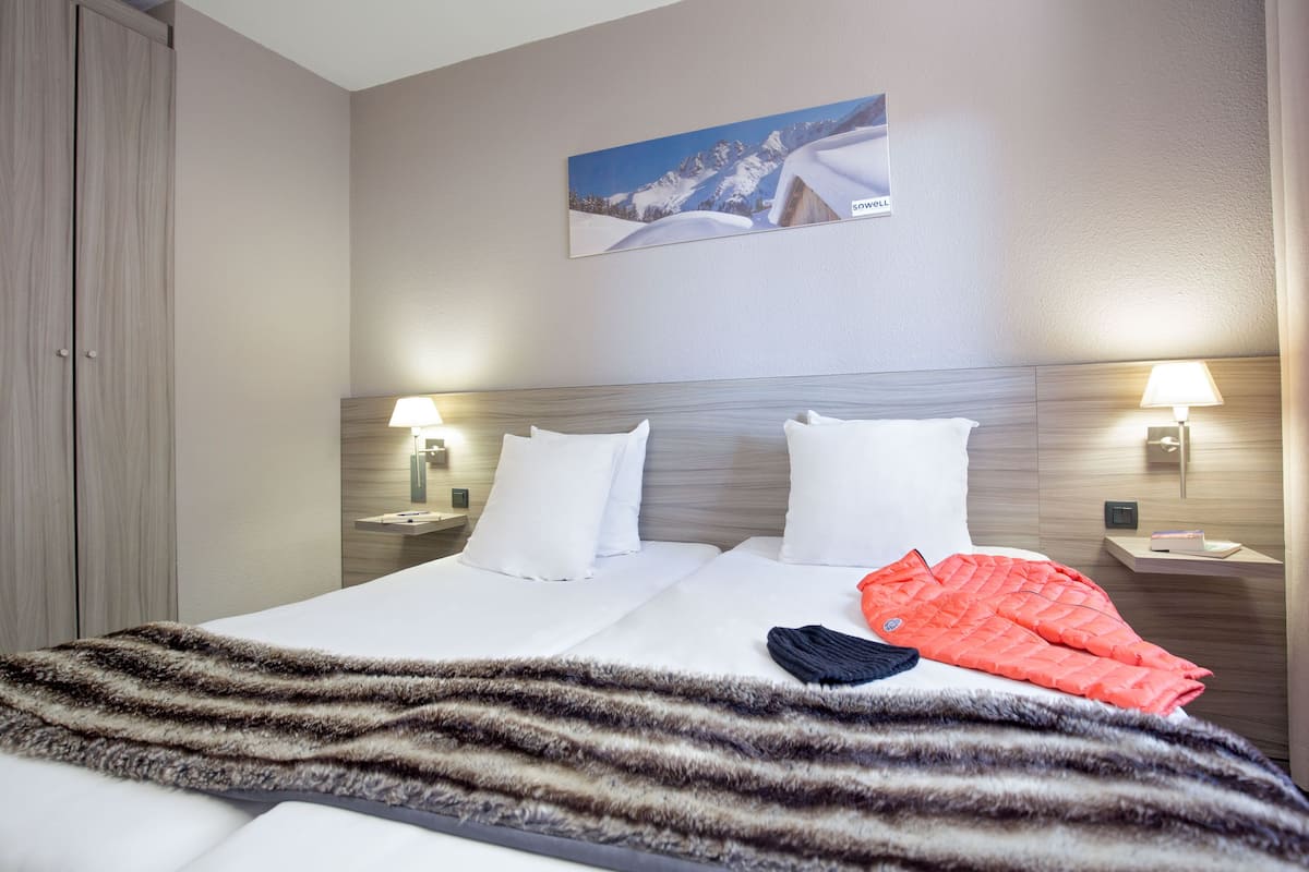 Une chambre d’hôtel comprenant un lit, deux tables de chevet, une petite table, une lampe fixée au mur et un tableau représentant une montagne enneigée.