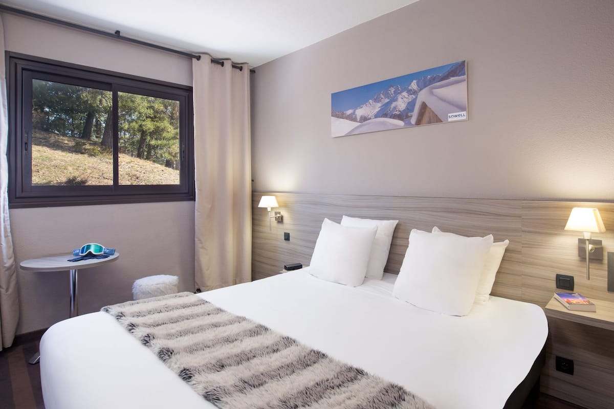 Une chambre d’hôtel avec un grand lit, une petite table sur laquelle reposent une paire de lunettes de soleil, une table de chevet avec une lampe et un tableau accroché au mur.