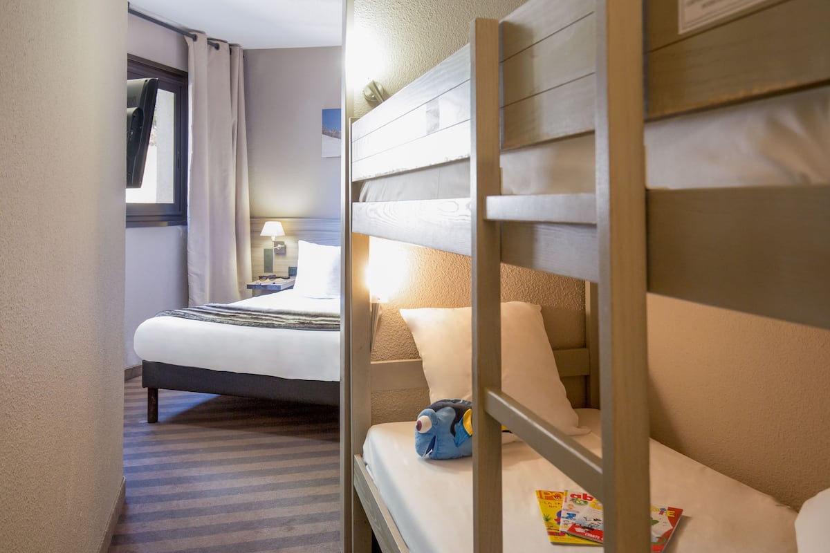 Une chambre d’hôtel avec un lit superposé, une fenêtre avec des rideaux, une table de chevet avec une lampe et une étagère fixée au mur.