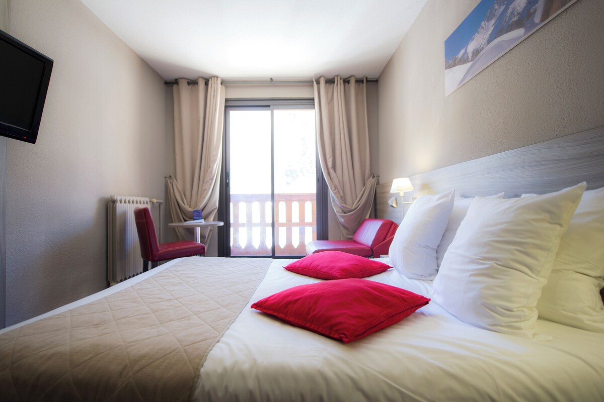 A hotel room with a bed, red and white pillows, a red chair, a table with a lamp, and a view of the outdoors through a window with curtains.