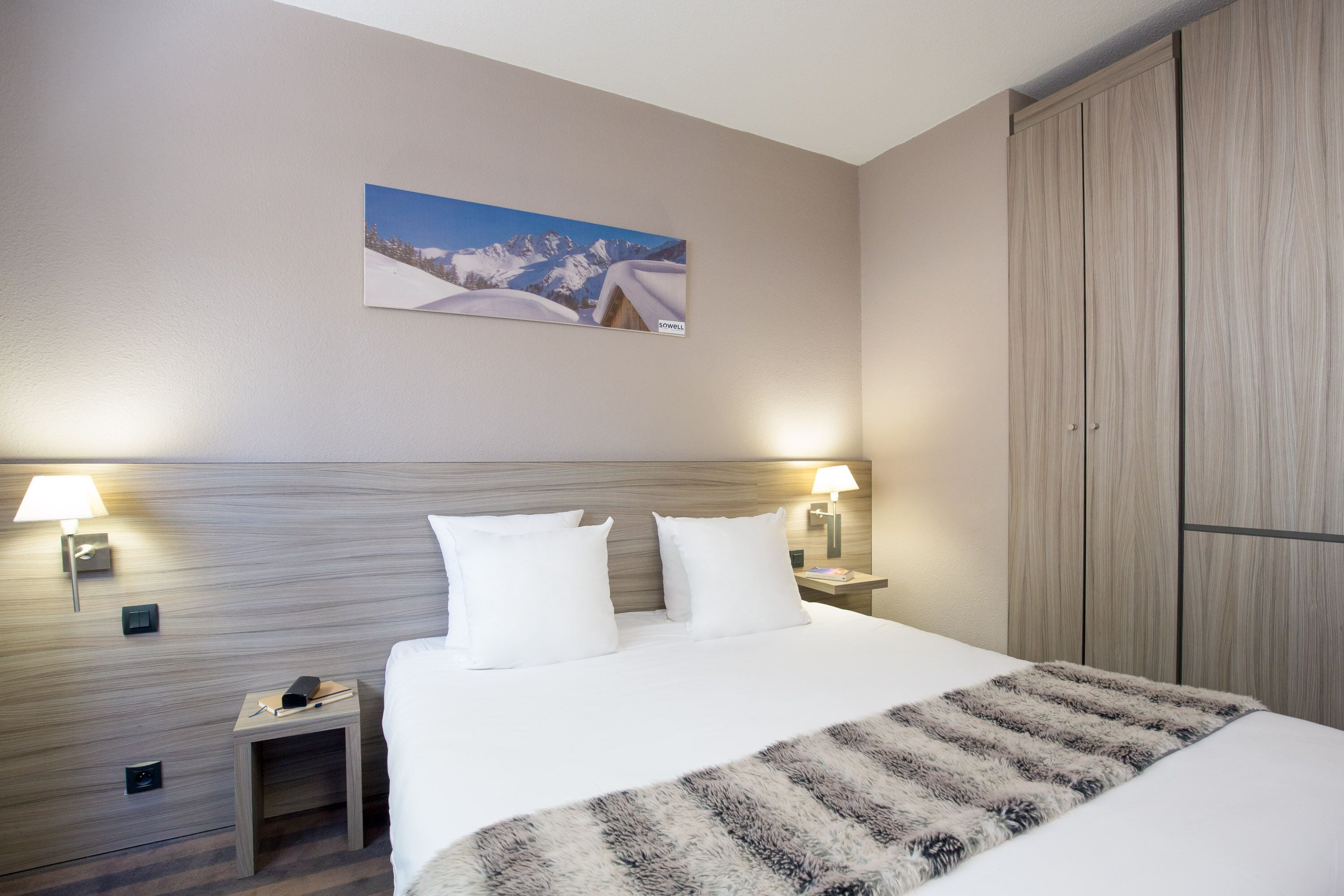 Hotelkamer met een groot bed, twee bedlampjes, een nachtkastje met telefoon en afstandsbediening, een berglandschap schilderij en een garderobe.