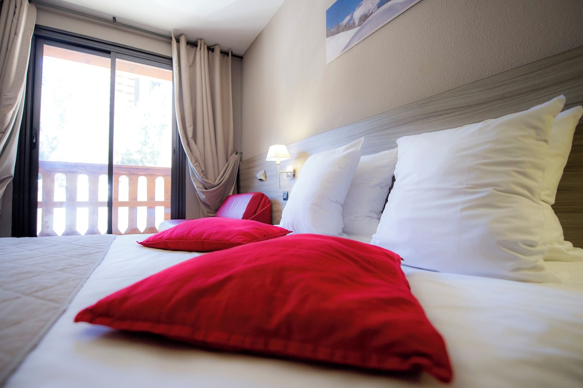 Une chambre d’hôtel avec un lit, des oreillers rouges, une table de chevet, une lampe et une vue sur un balcon.
