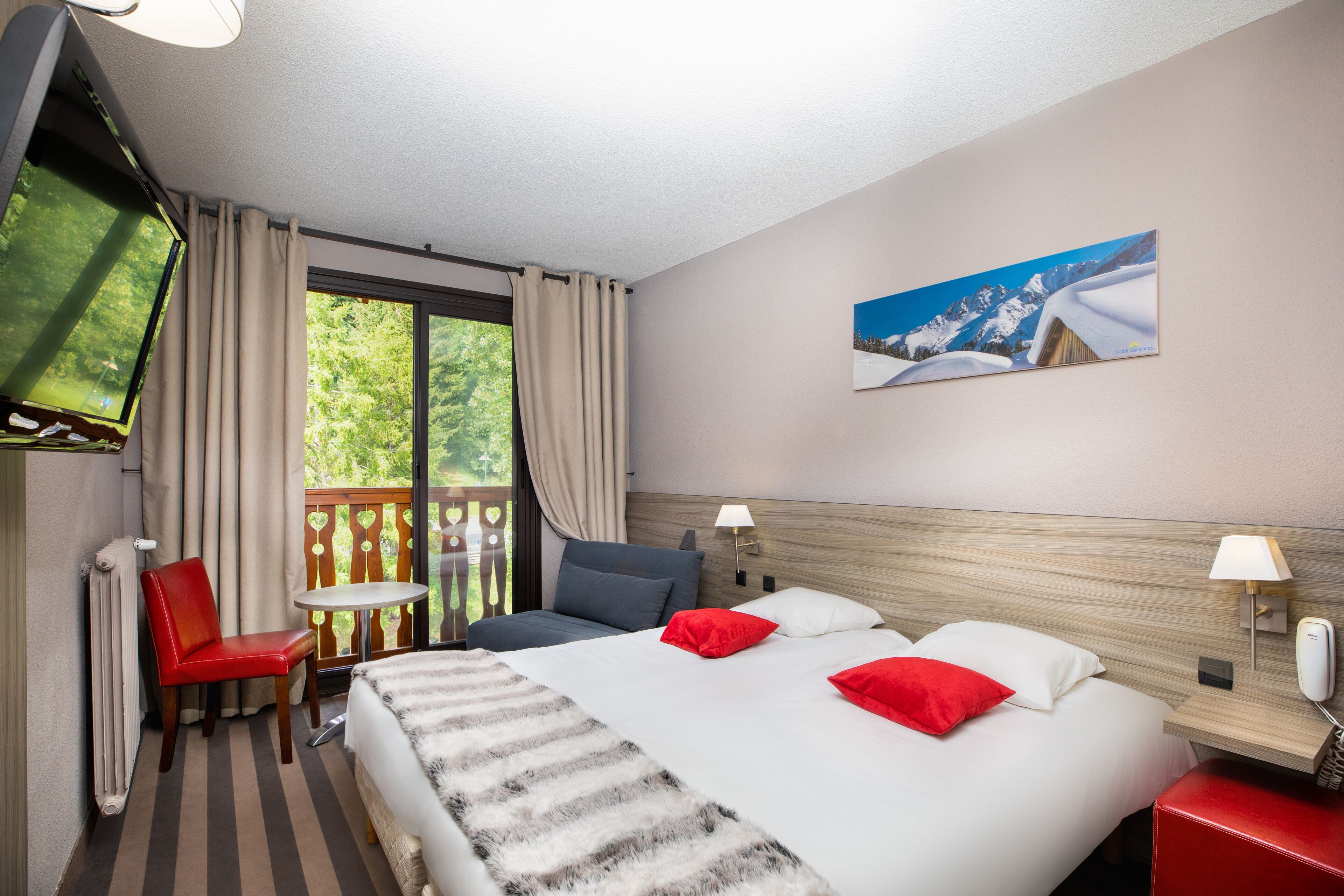 Habitación de hotel con cama, televisión, una silla, una mesita y ventana con cortinas.