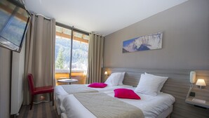 Habitación de hotel con dos camas, una silla roja, una mesita, un cuadro en la pared y una ventana con vistas a la montaña.
