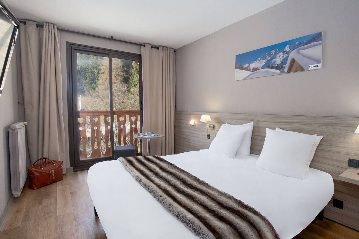 Une chambre d’hôtel avec un grand lit, une tête de lit en bois, une table de chevet avec une lampe, une vue sur l’extérieur grâce à une porte coulissante et un tableau au mur.