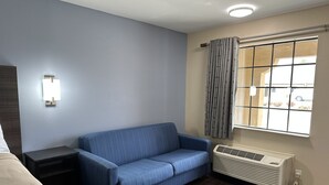 A hotel room with a blue sofa, a bedside table, a lamp, a window with curtains, and a radiator. - Express Inn & Suites (Hartselle)