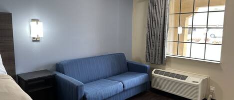 Une chambre d’hôtel avec un canapé bleu, une table de chevet, une lampe, une fenêtre avec des rideaux et un radiateur.
