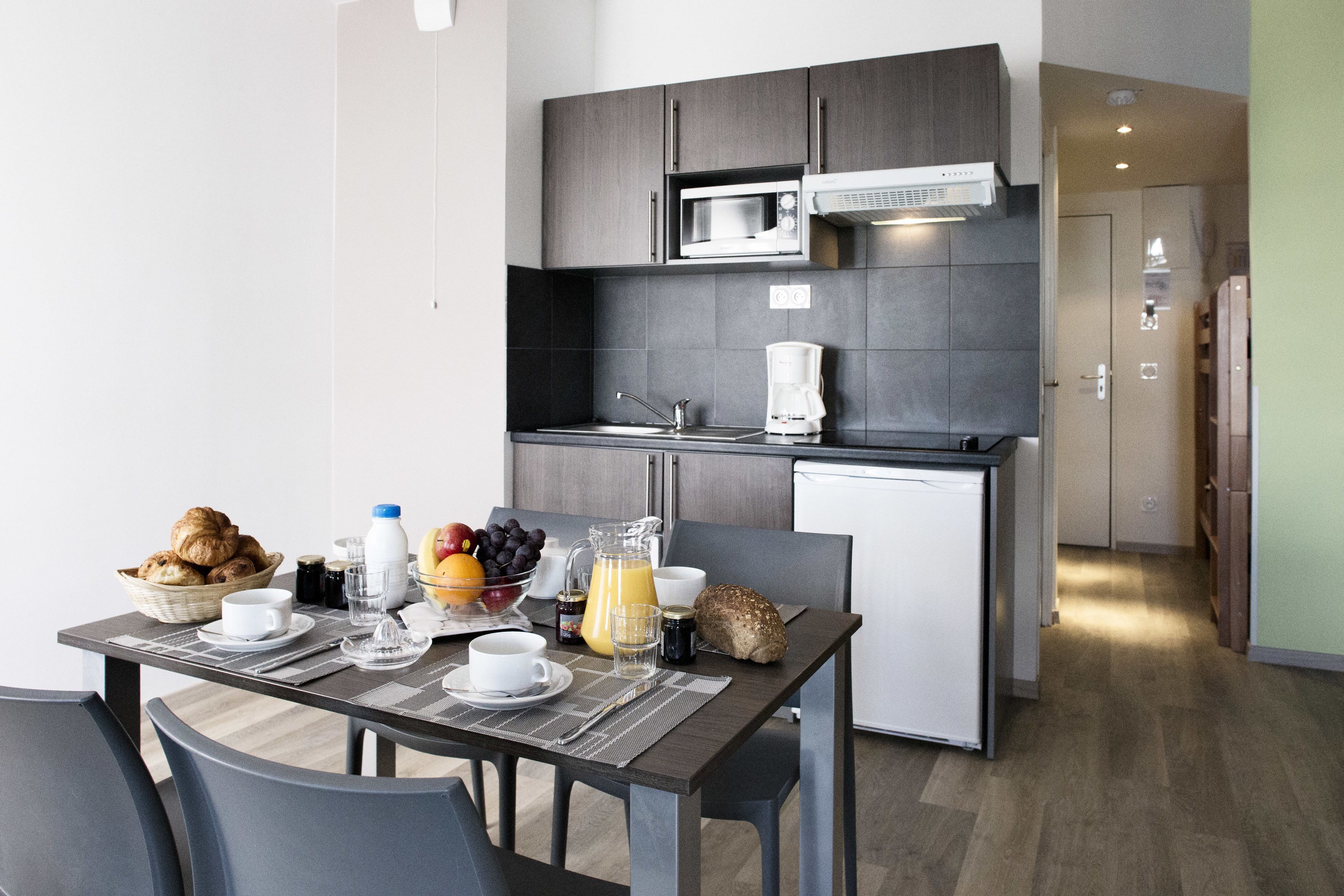 A modern kitchen with a microwave, coffee maker, and a small refrigerator. There is a dining table set with plates, cups, and a pitcher of orange juice.
