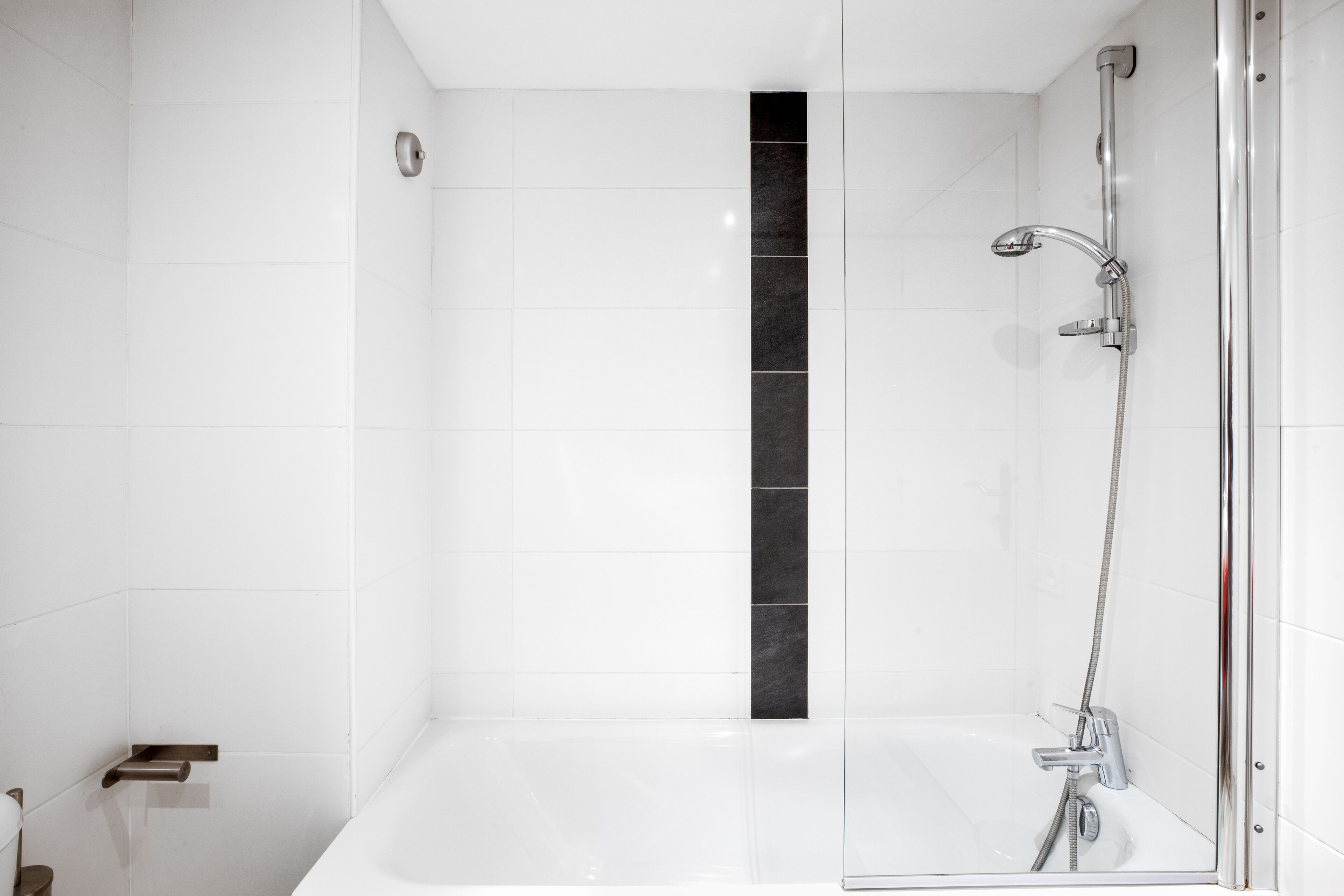 Un baño moderno con azulejos blancos, un detalle vertical negro, ducha con mampara de vidrio y grifo cromado.