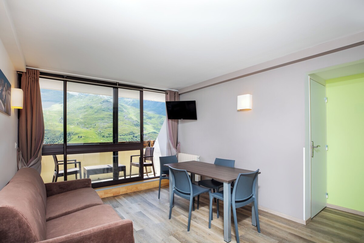 A hotel room with a large window, a dining table with chairs, a sofa, and a television mounted on the wall.