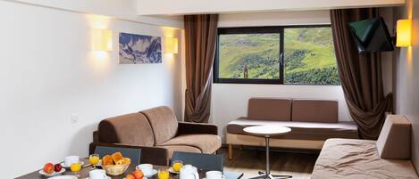 Ein modernes Hotelzimmer mit Essbereich, einer Couch, einem Fernseher und einem großen Fenster mit Blick auf eine grüne Landschaft.