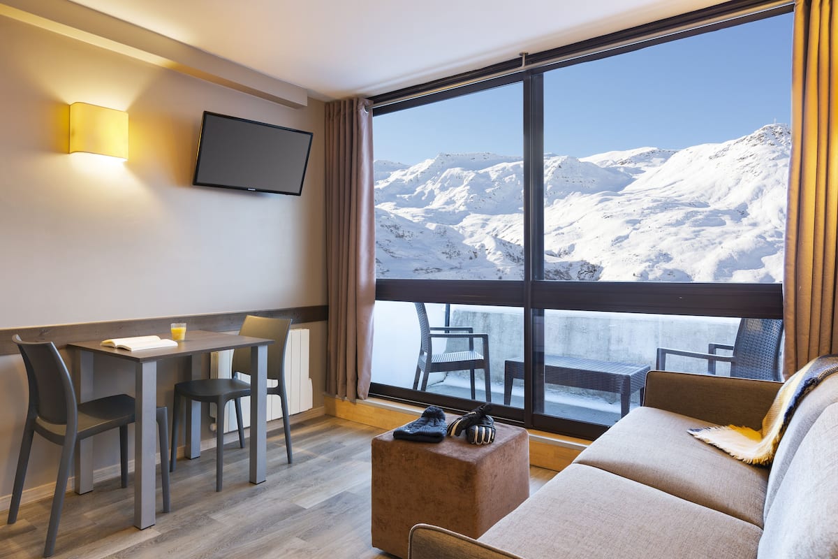 A modern hotel room with a large window offering a mountain view, a dining area with a table and chairs, and a cozy seating area with a sofa and ottoman.