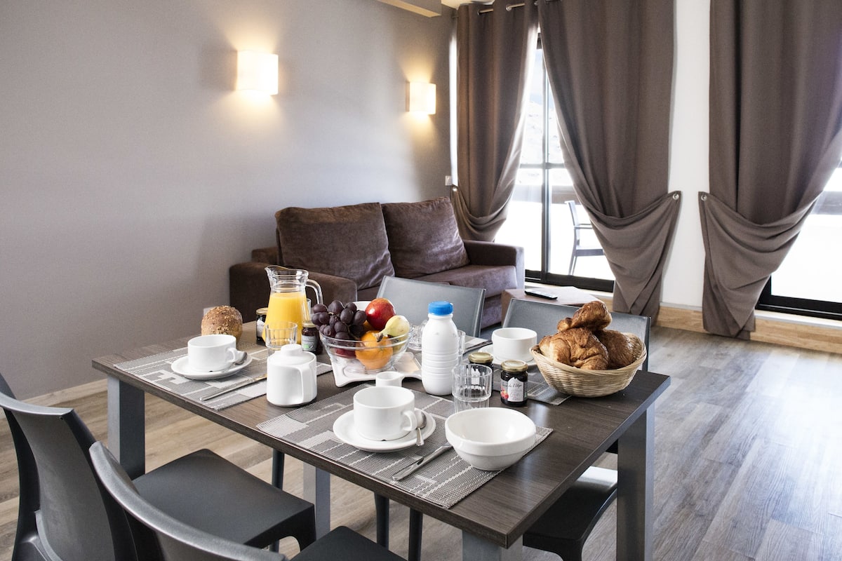 A breakfast setup with a table, chairs, breakfast items, and a sofa in a modern room with curtains and a window.