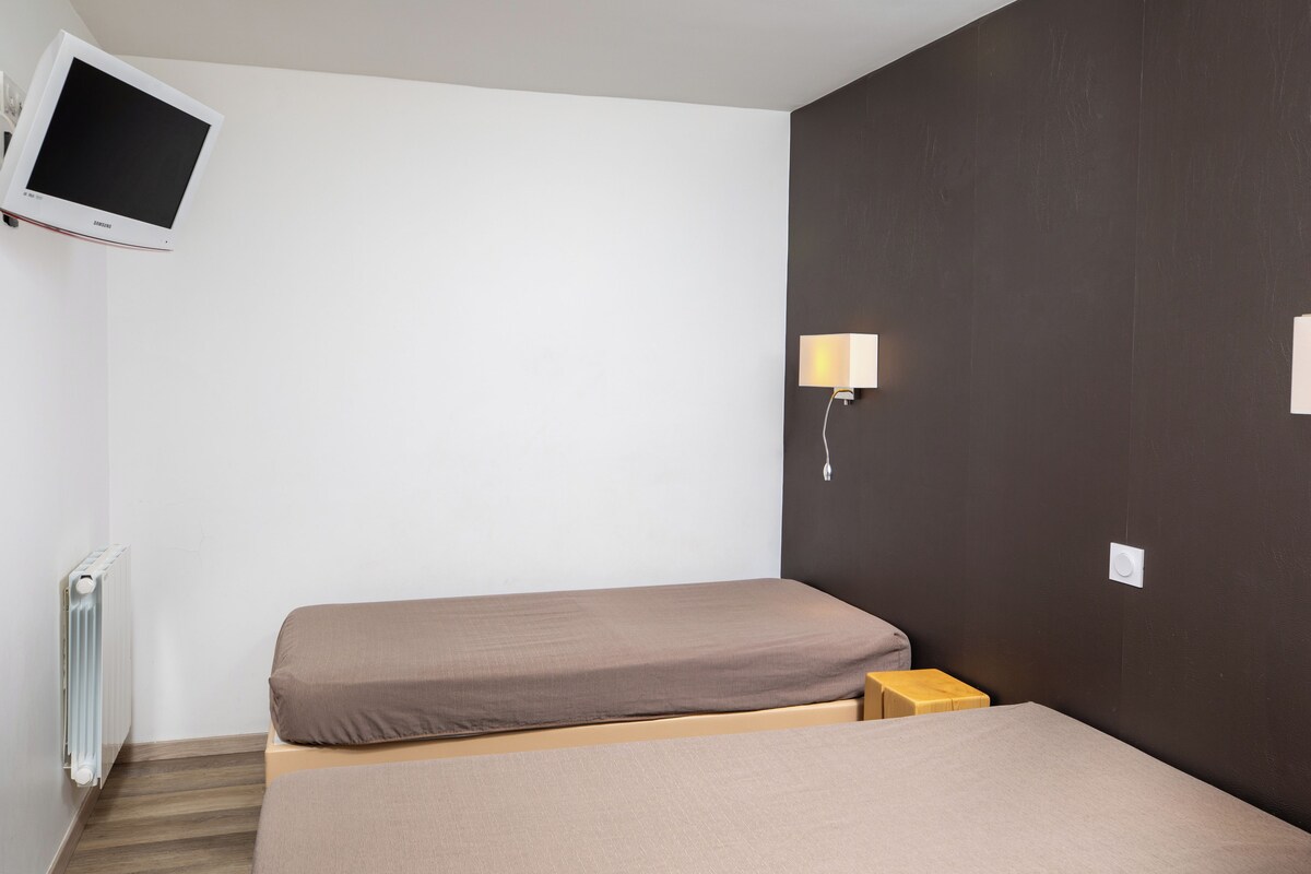 A small, simple room with two beds, a wall-mounted TV, and a bedside table with a lamp.