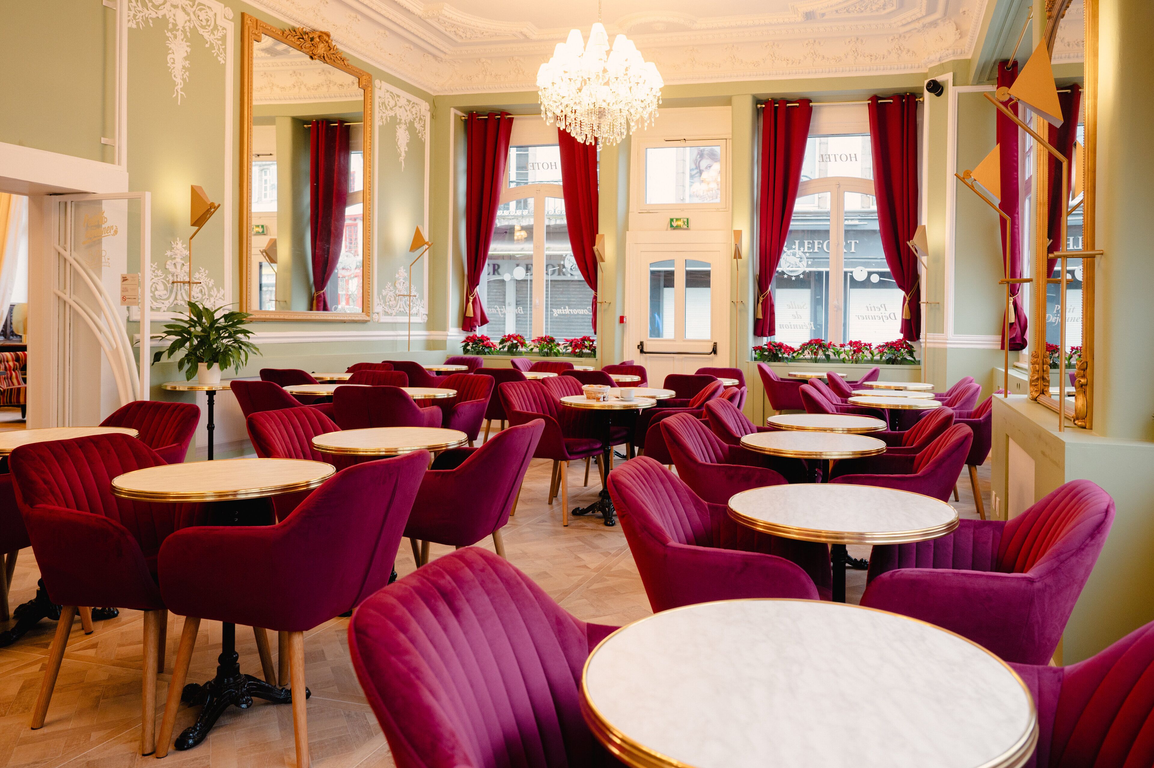 Un restaurant avec des fauteuils en velours rouge et des tables rondes, de grandes fenêtres ornées de rideaux rouges et un lustre.