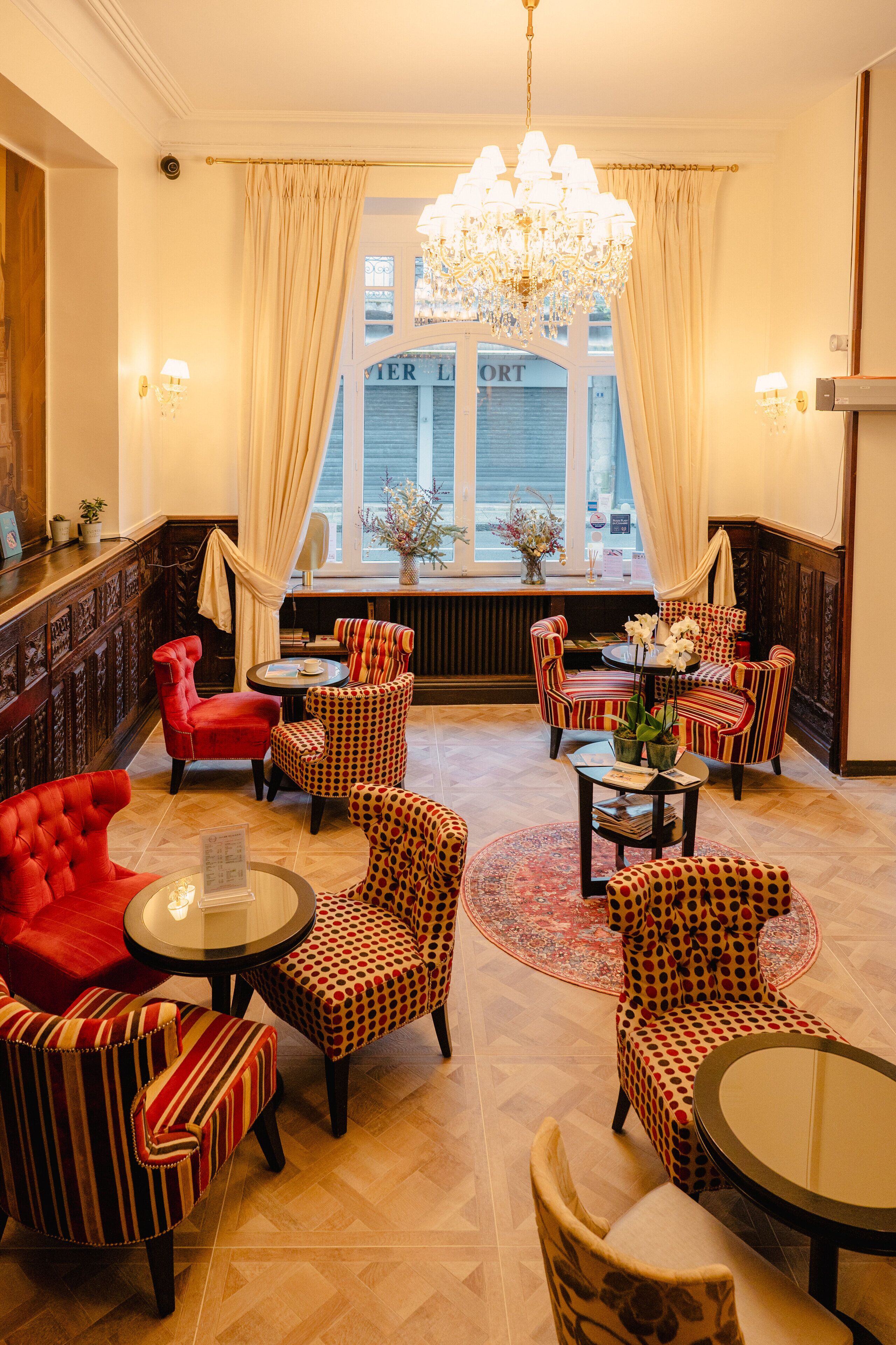 A hotel lobby with a chandelier, striped armchairs, a red armchair, and a view of the street through a window.