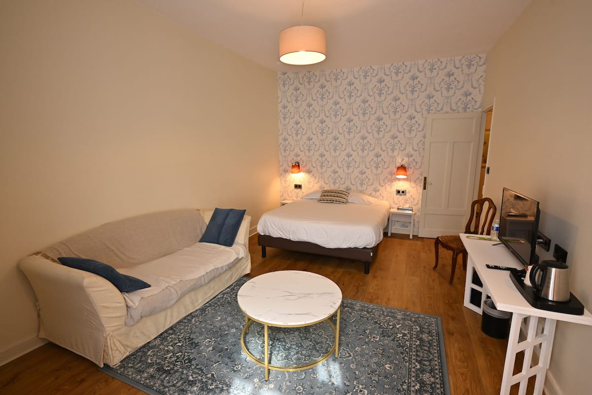 Une petite pièce bien éclairée, avec un canapé, une table basse ronde, un lit, un bureau avec un ordinateur et un tapis à motifs.