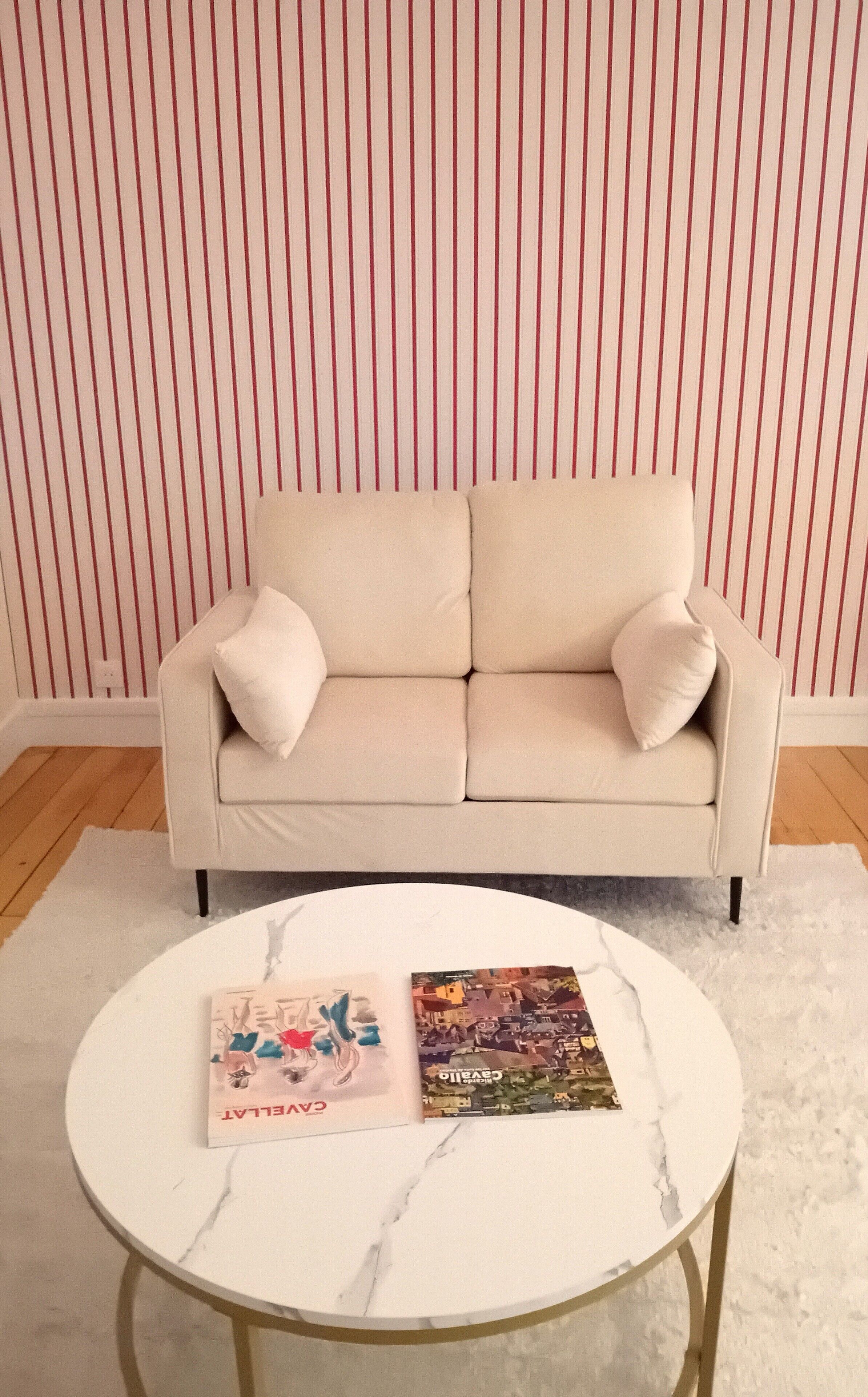 A beige sofa with two pillows against a striped wallpaper backdrop. In front of the sofa is a white marble coffee table with two books.