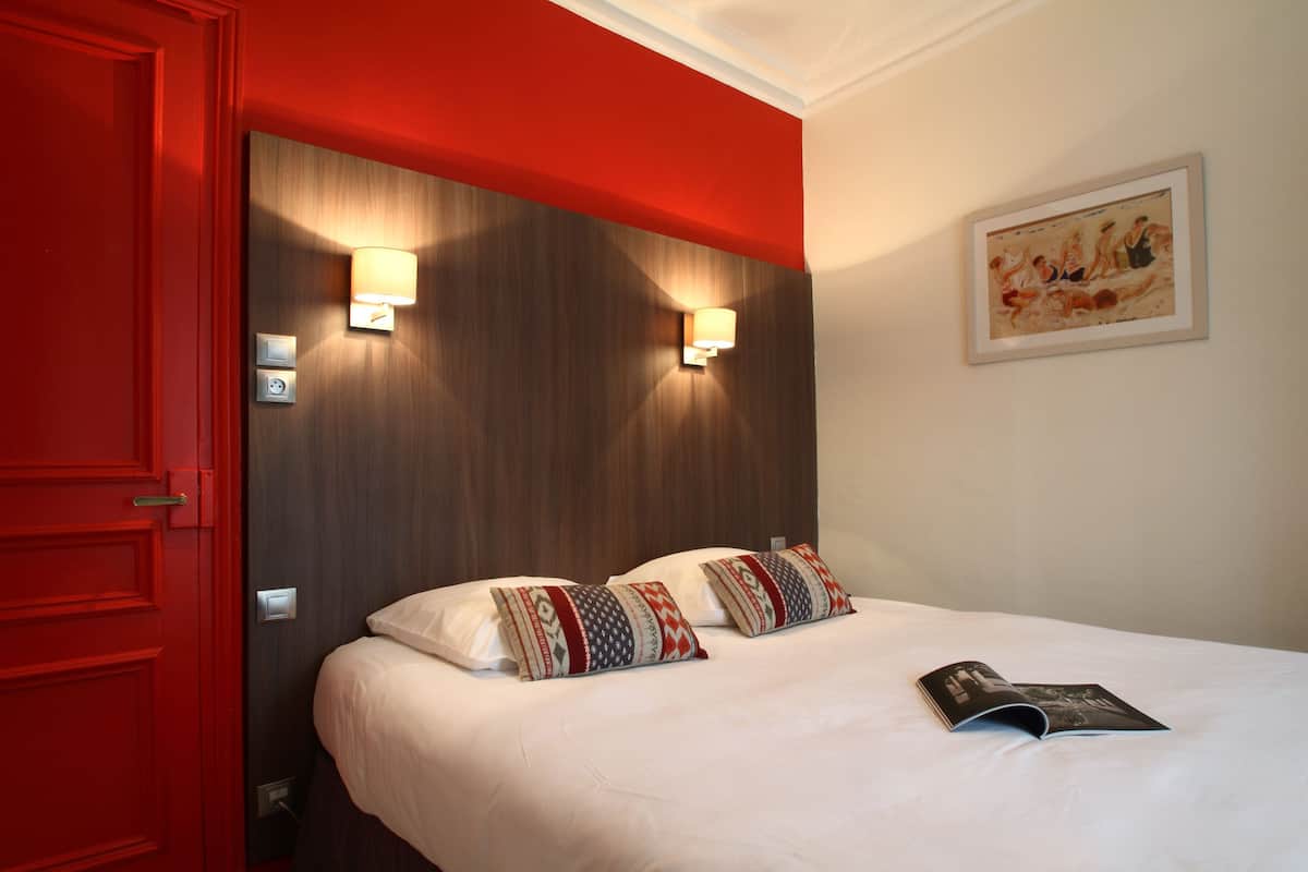 Une chambre d’hôtel avec une porte rouge, deux lampes de chevet, un lit avec des oreillers à motifs et un tableau encadré au mur.