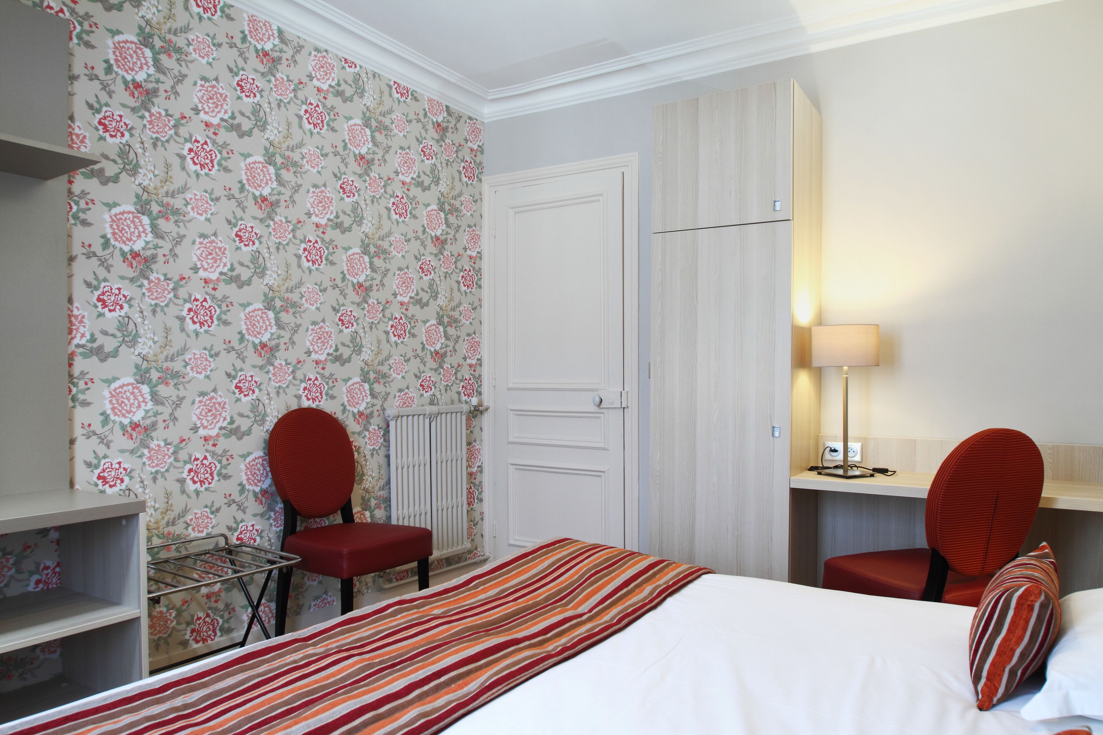 Habitación de hotel con una cama, dos sillas rojas, un escritorio con lámpara y papel tapiz con motivos florales.