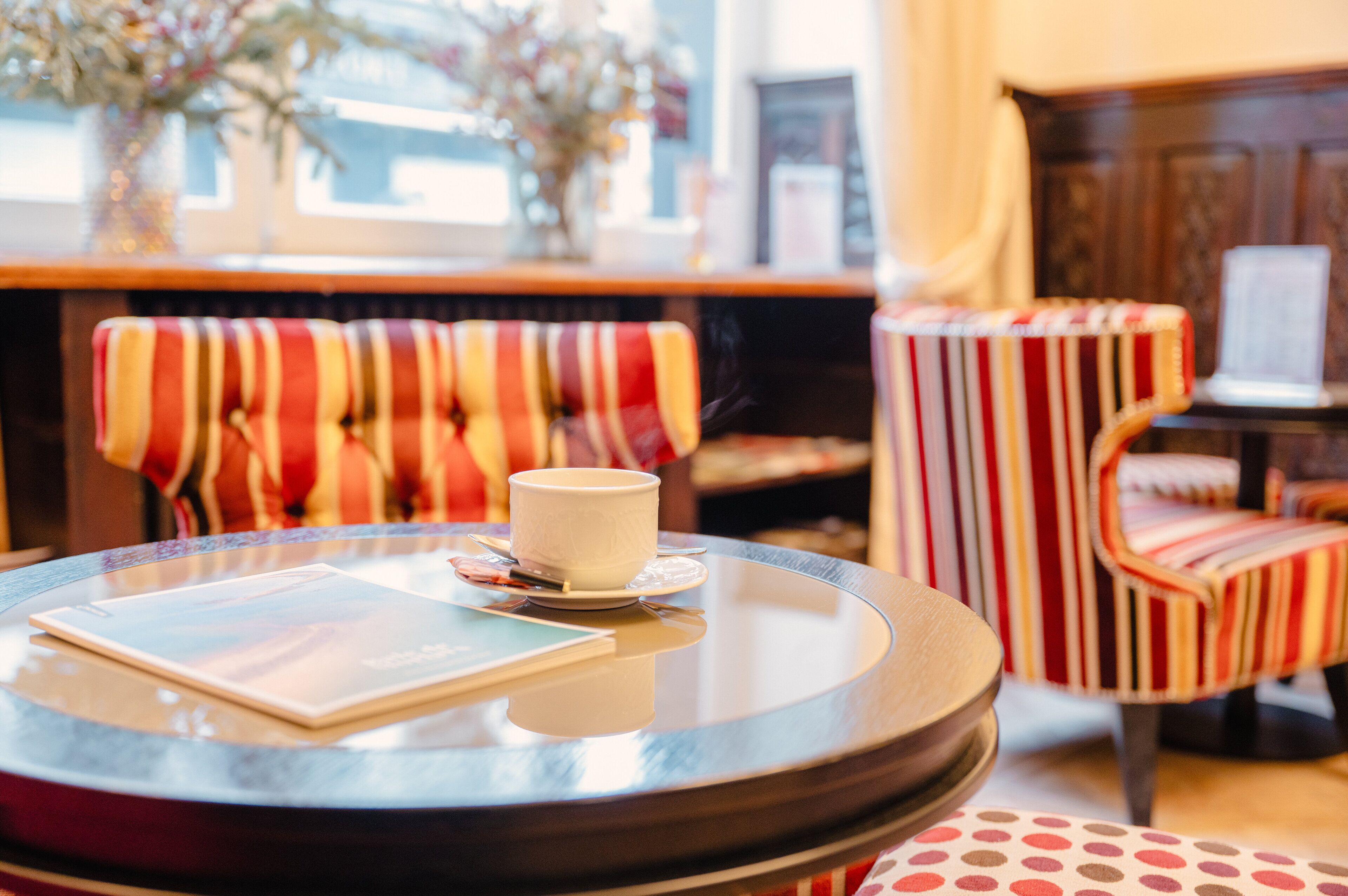A round table with a white cup and a menu, a striped armchair, and a wooden cabinet in a cozy room.