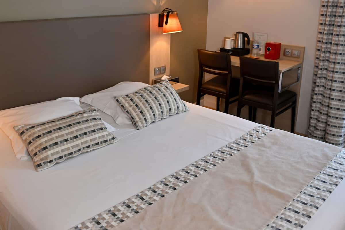 Une chambre d’hôtel avec un grand lit, deux tables de chevet, un bureau avec une chaise, une lampe fixée au mur et un rideau.