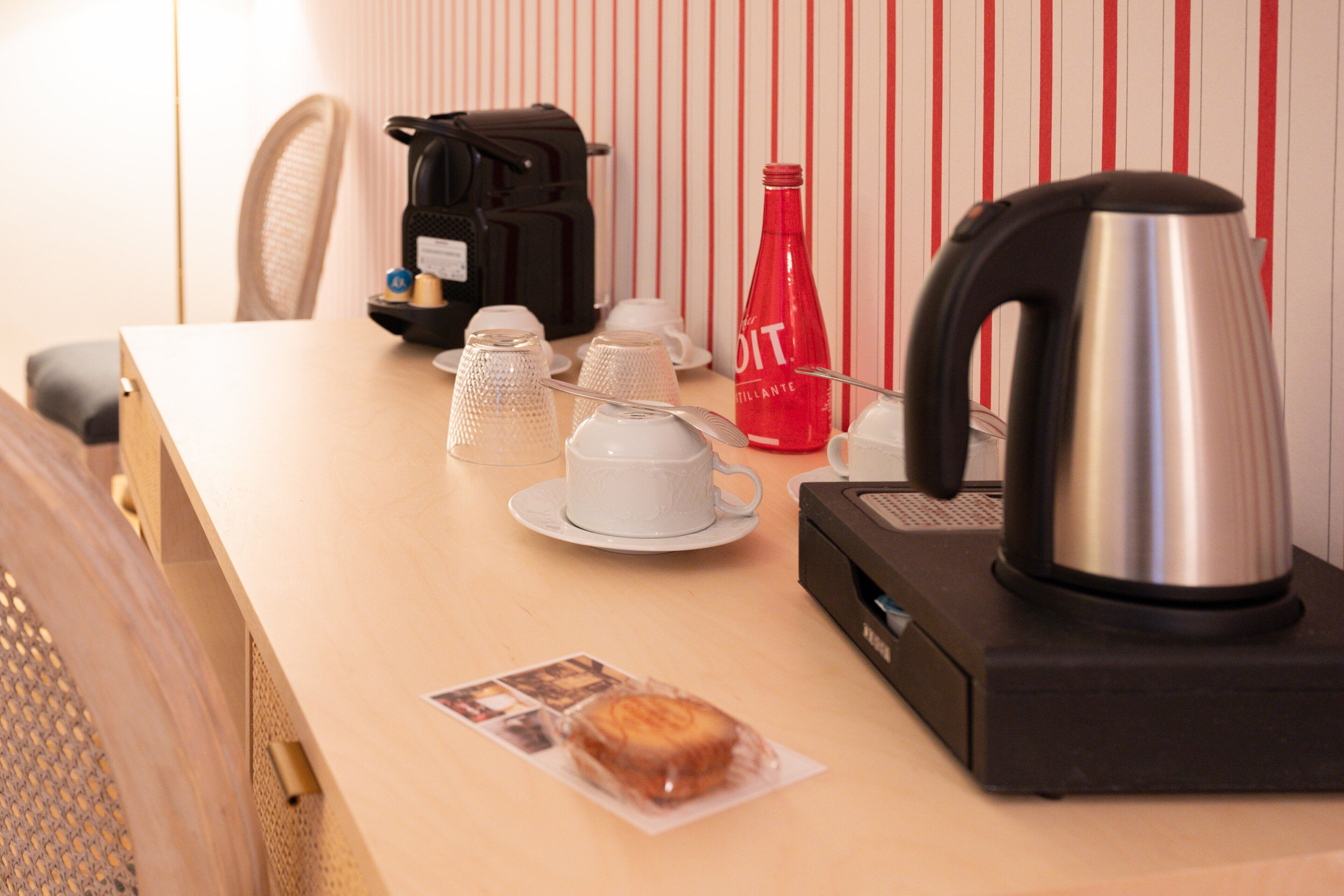 A hotel room with a wooden table, a coffee maker, a red bottle, a kettle, and a wrapped pastry.