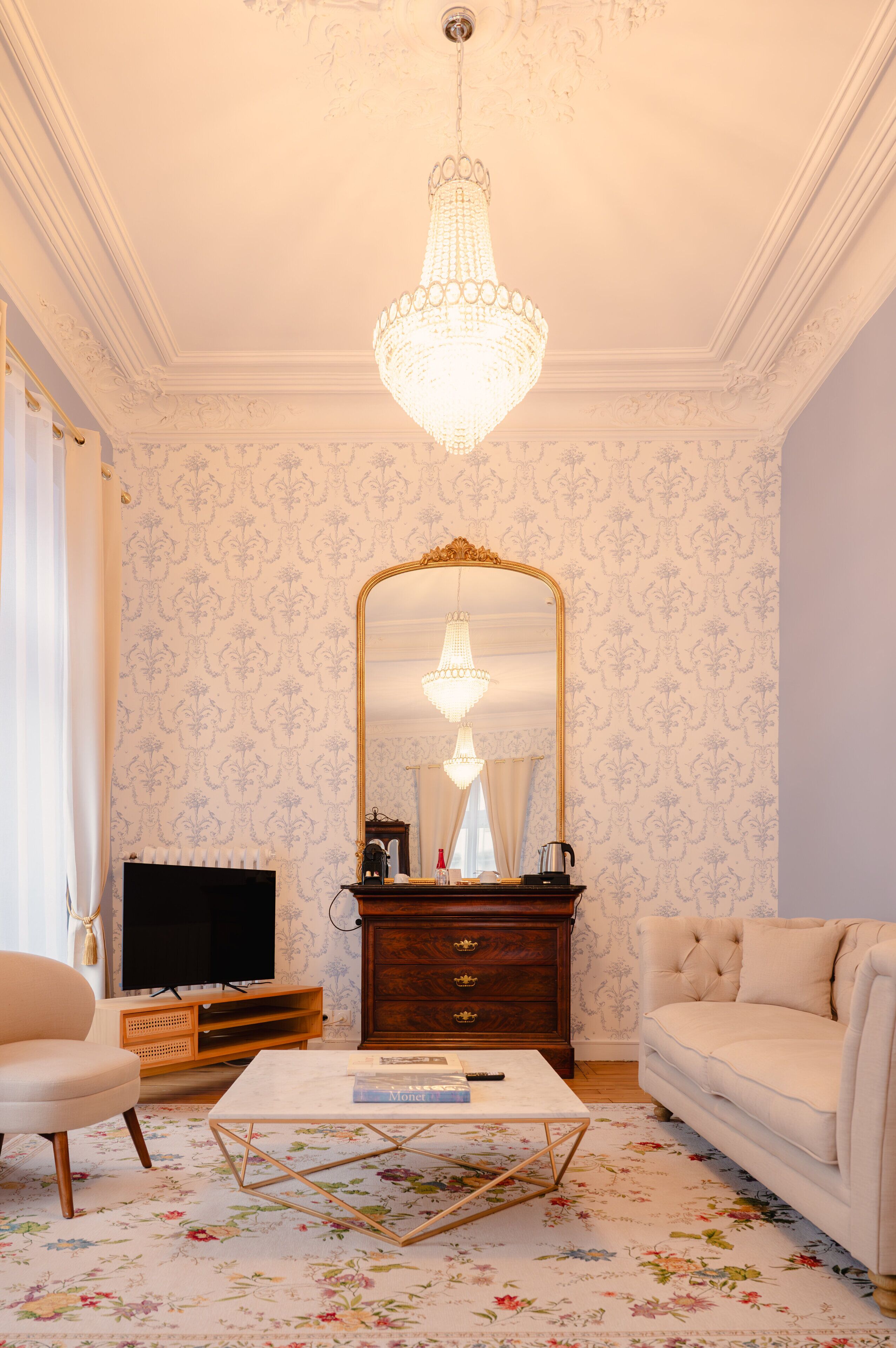 Ein Wohnzimmer mit einer Chandelüster, einem großen Spiegel, einem Fernseher, einem Couchtisch und einem Sofa.