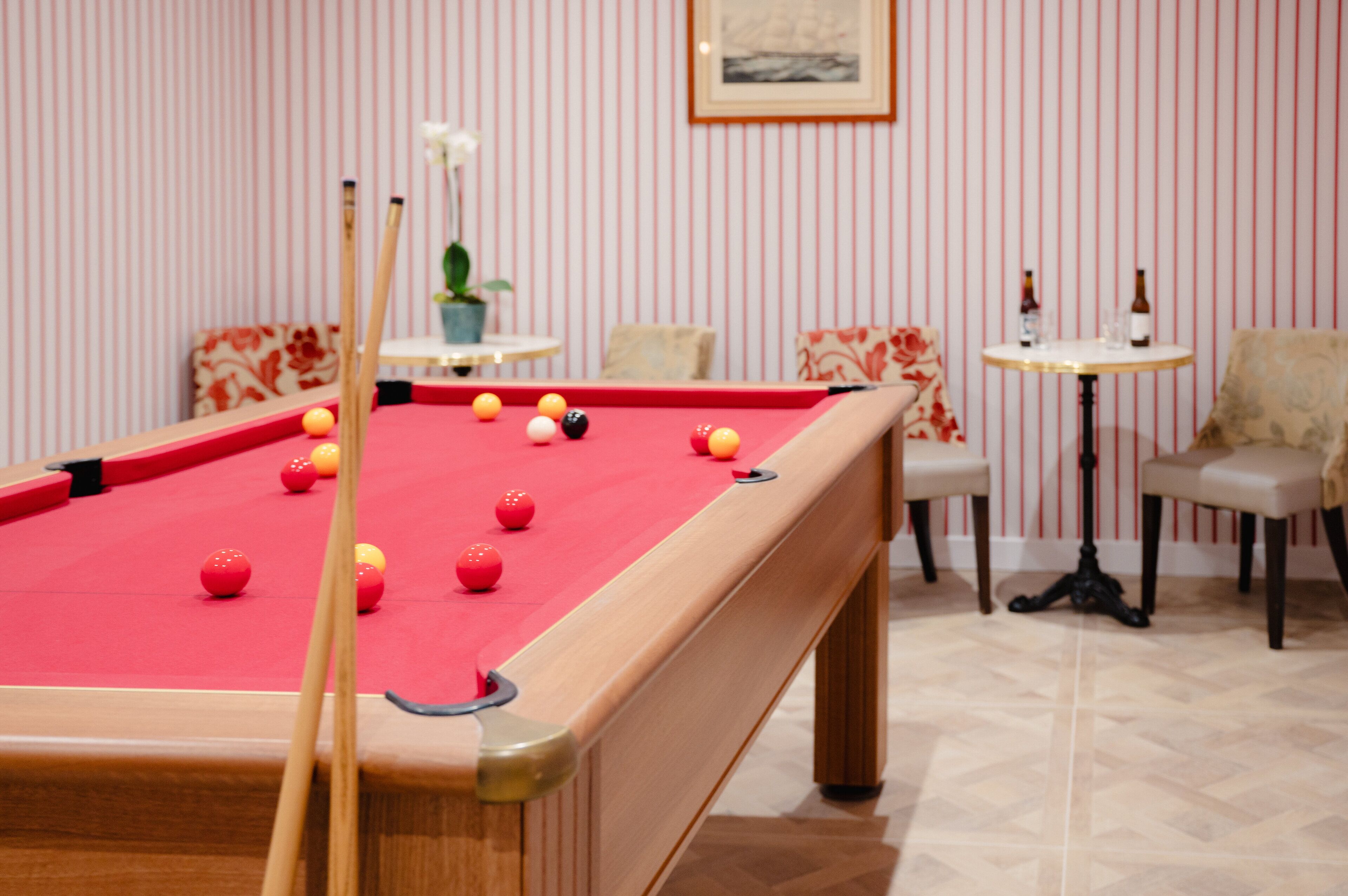A pool table with balls, a chair, a small table with bottles, and striped wallpaper.