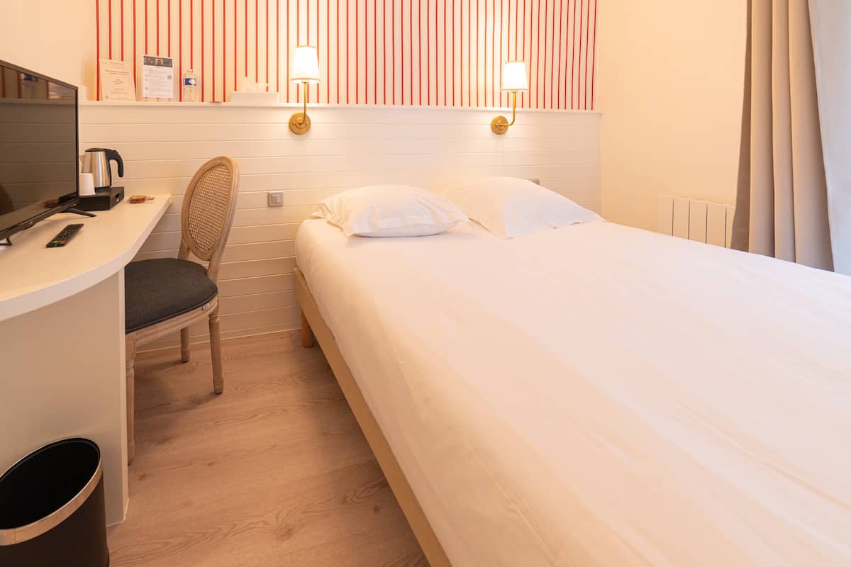 Une chambre d’hôtel comprenant un lit, un bureau avec une chaise, une télévision et une étagère fixée au mur, sur laquelle sont posés divers objets.
