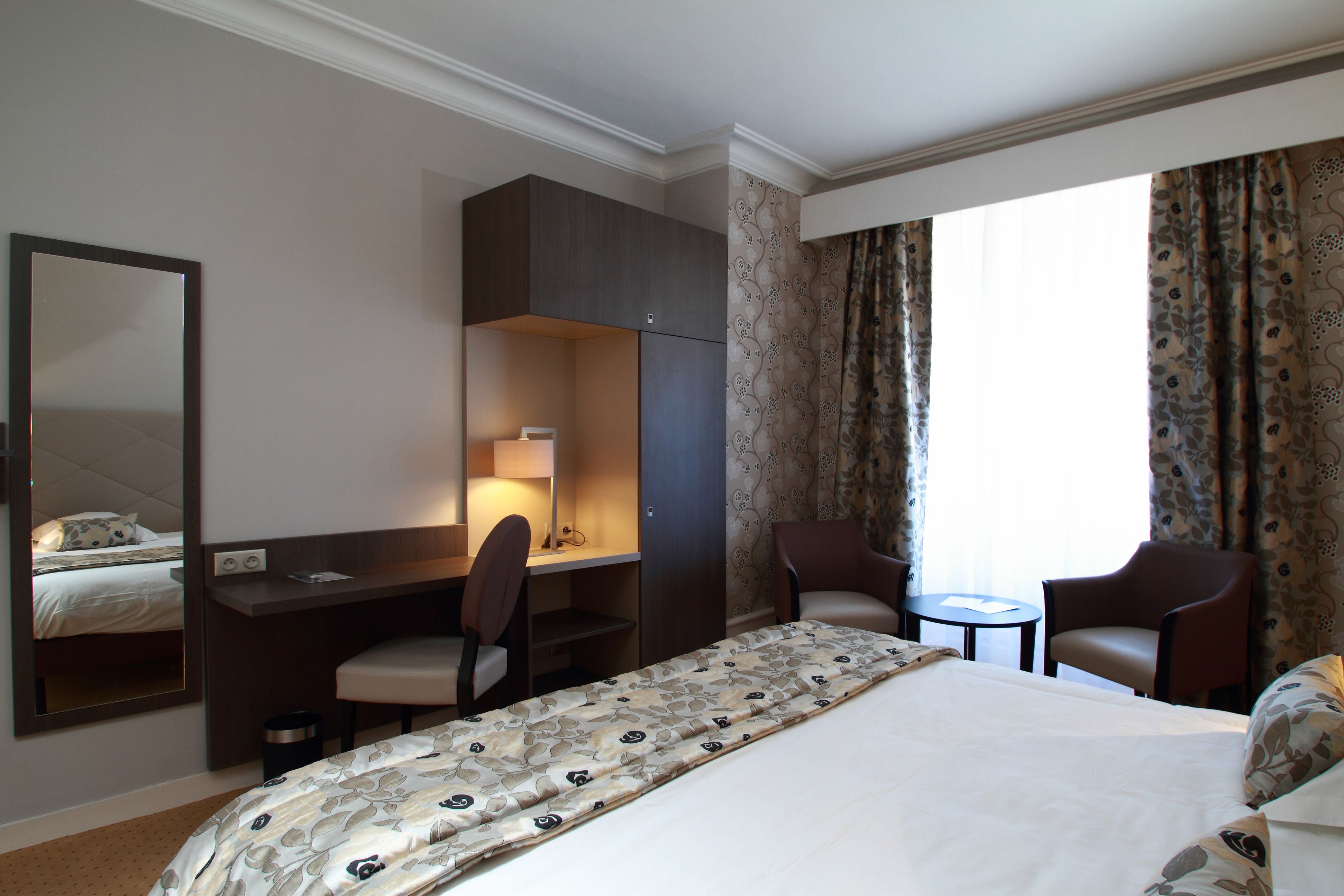 Habitación de hotel con cama, escritorio, dos sillas, una mesita, un espejo y ventana con cortinas.