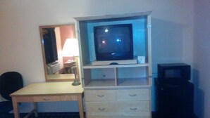 A hotel room with a desk, a television on a stand, a mirror, and a small cabinet.