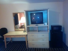 Habitación de hotel con escritorio, televisor sobre un soporte, espejo y un mueble pequeño.