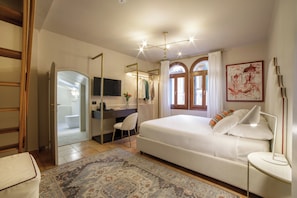 Quarto com cama, escrivaninha, cadeira, televisão e banheiro visível por uma porta aberta.