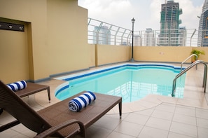 A rooftop pool with two wicker lounge chairs, a metal pool ladder, and a view of surrounding high-rise buildings. - Fernandina 88 Suites Hotel (QUEZON CITY)