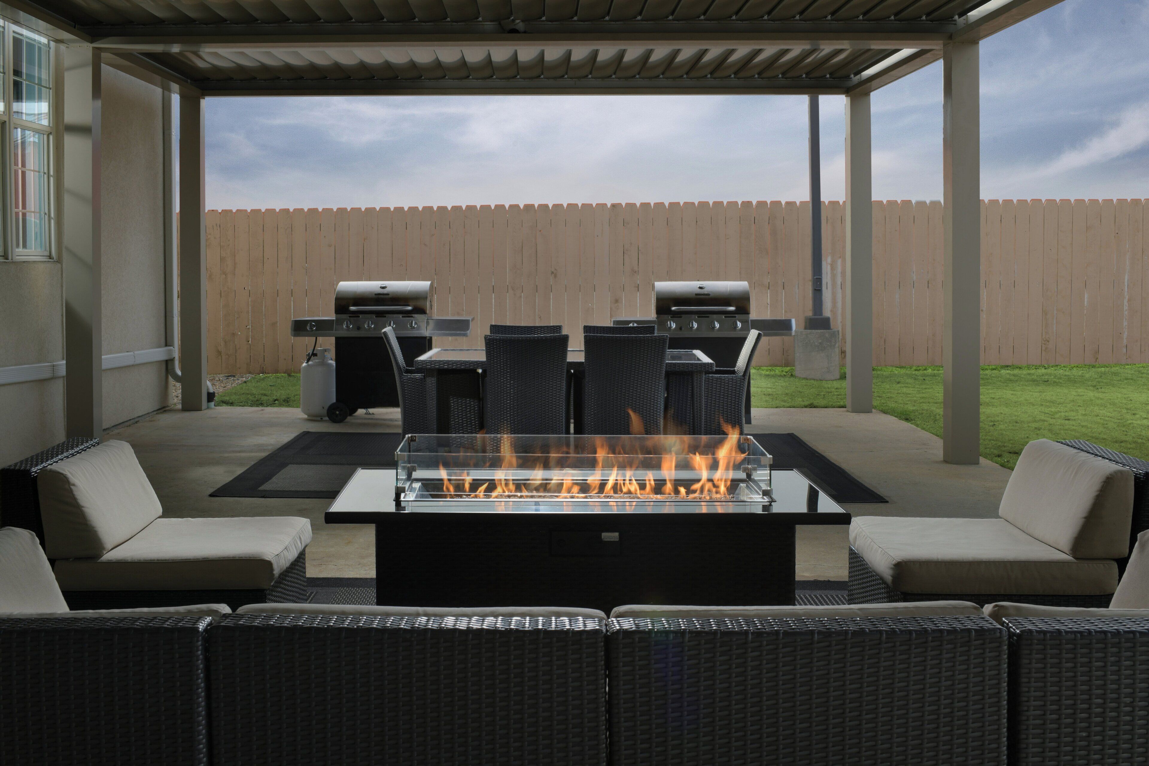 An outdoor patio with a fire feature, two barbecue grills, wicker furniture, and a wooden fence in the background.