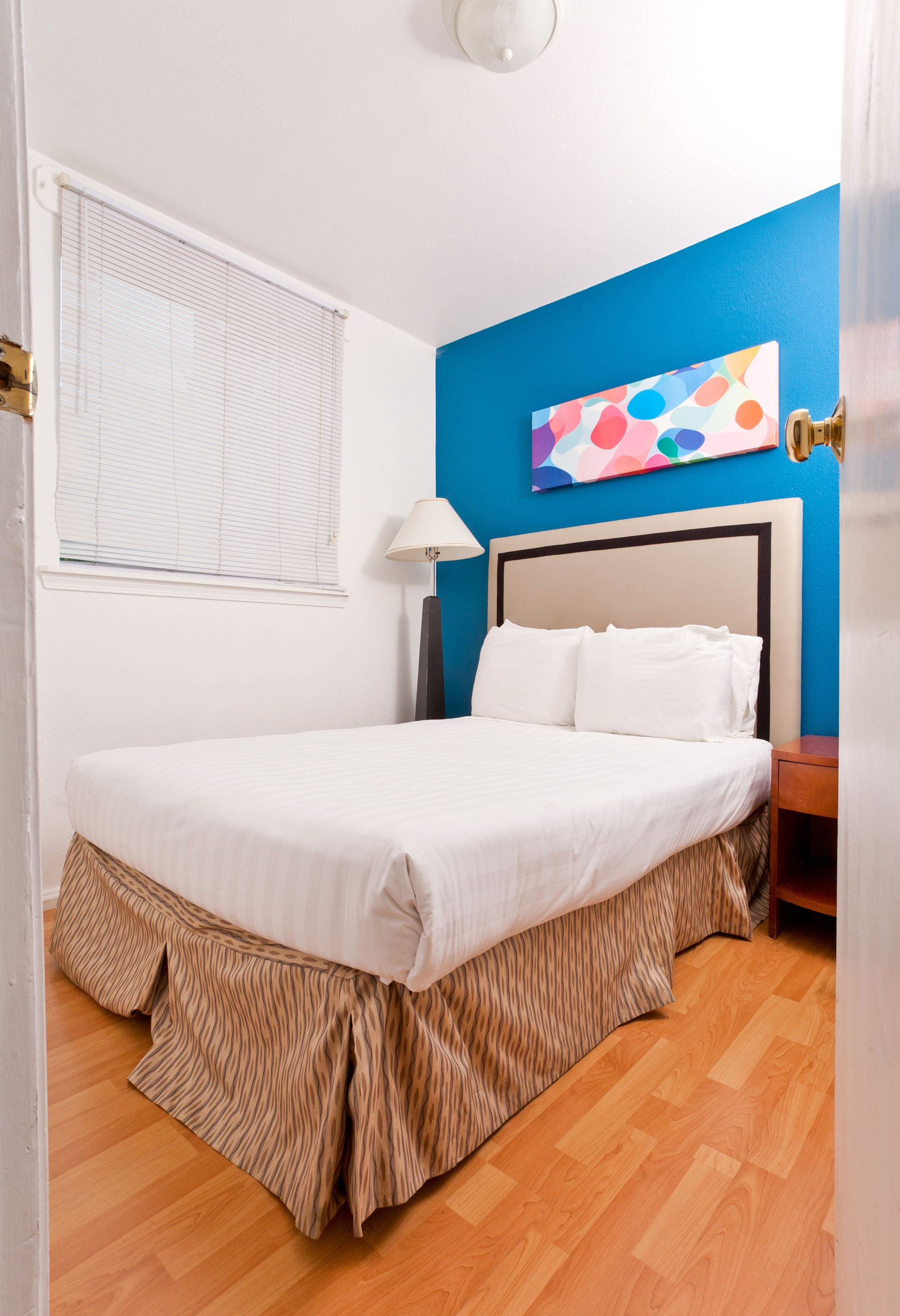 A bedroom with a bed, a window with blinds, a wall painting, and a lamp.