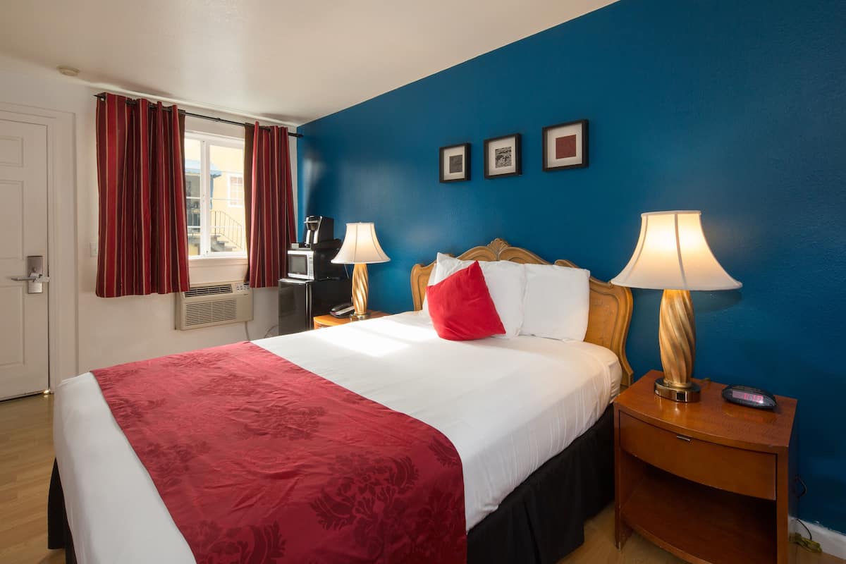 A hotel room with a bed, two bedside tables with lamps, a window with curtains, a wall with three framed pictures, and a door.