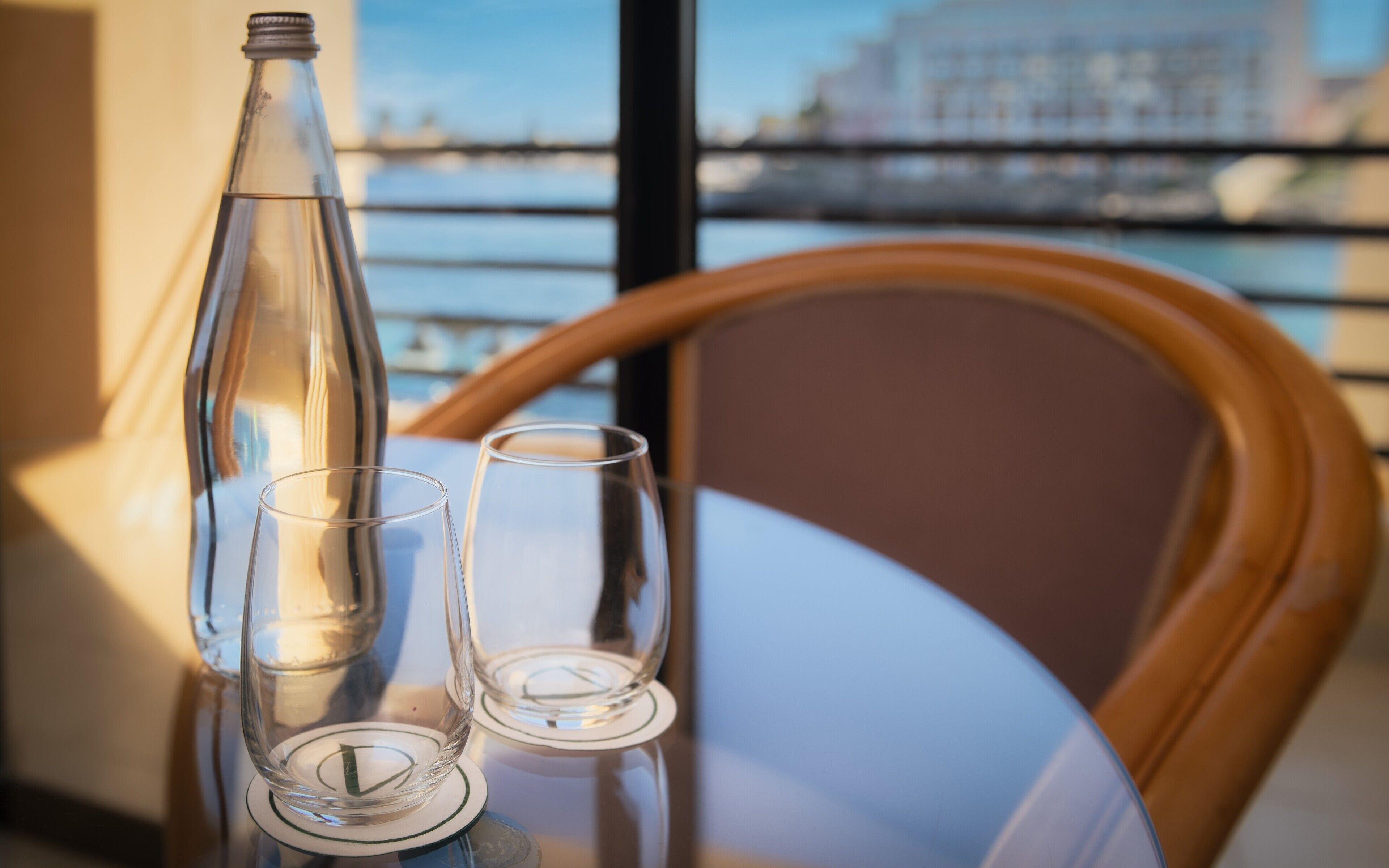 A table with a bottle and two glasses, a wooden chair, and a view of a bridge and buildings through a window.