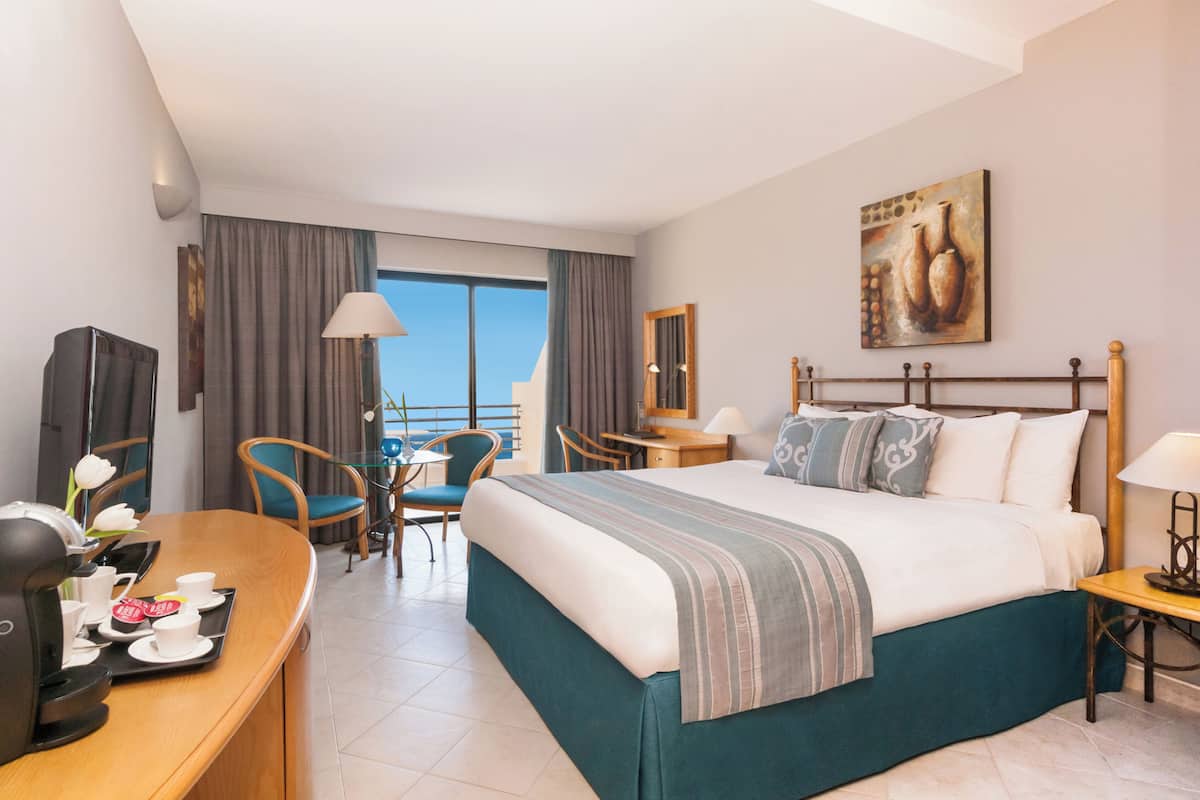 A hotel room with a bed, a desk with a coffee maker, a chair, a table, and a view of the outside.