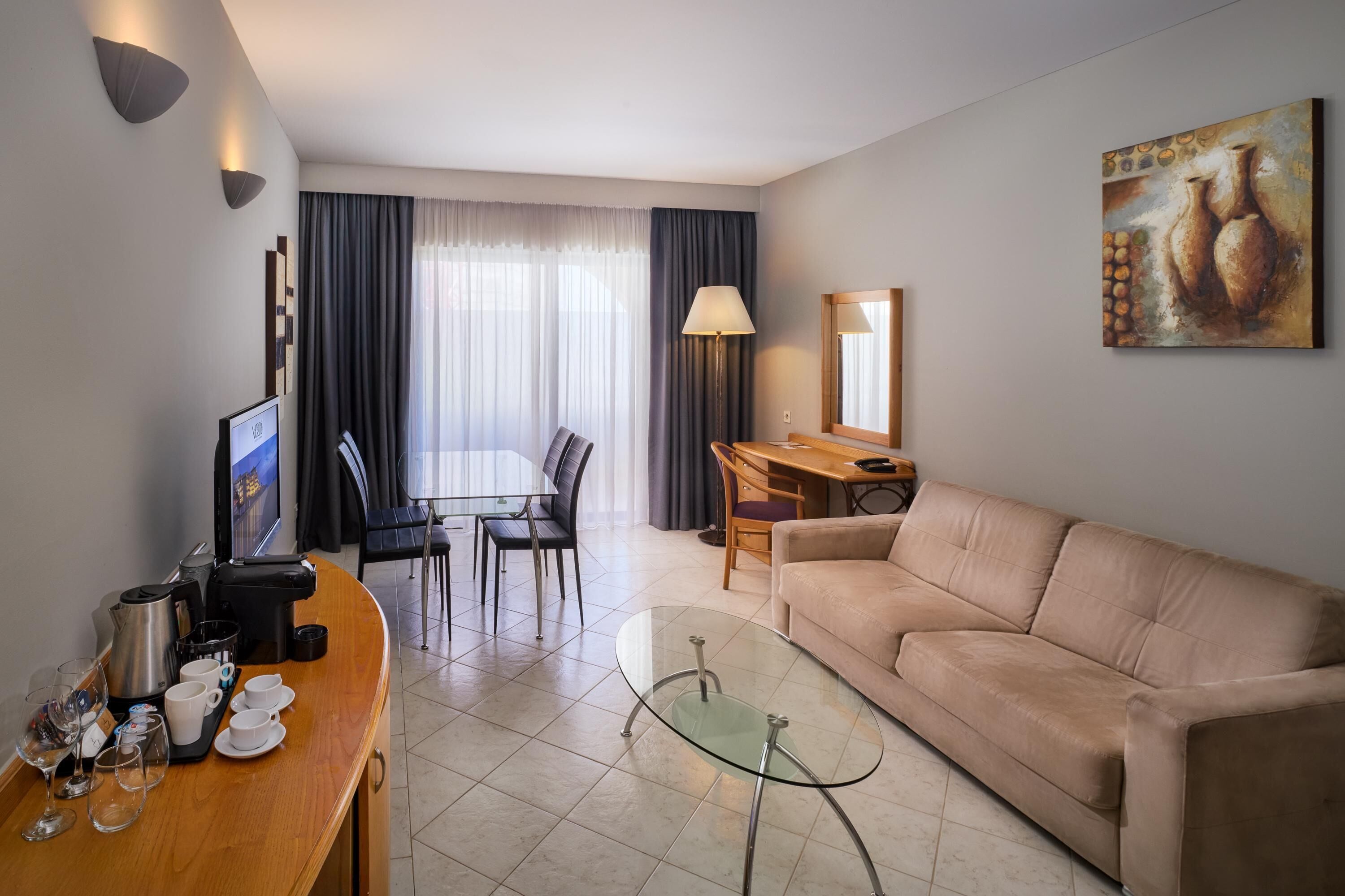 Habitación de hotel con sofá, una mesa de centro de vidrio, zona de comedor con sillas, televisor y un cuadro en la pared.