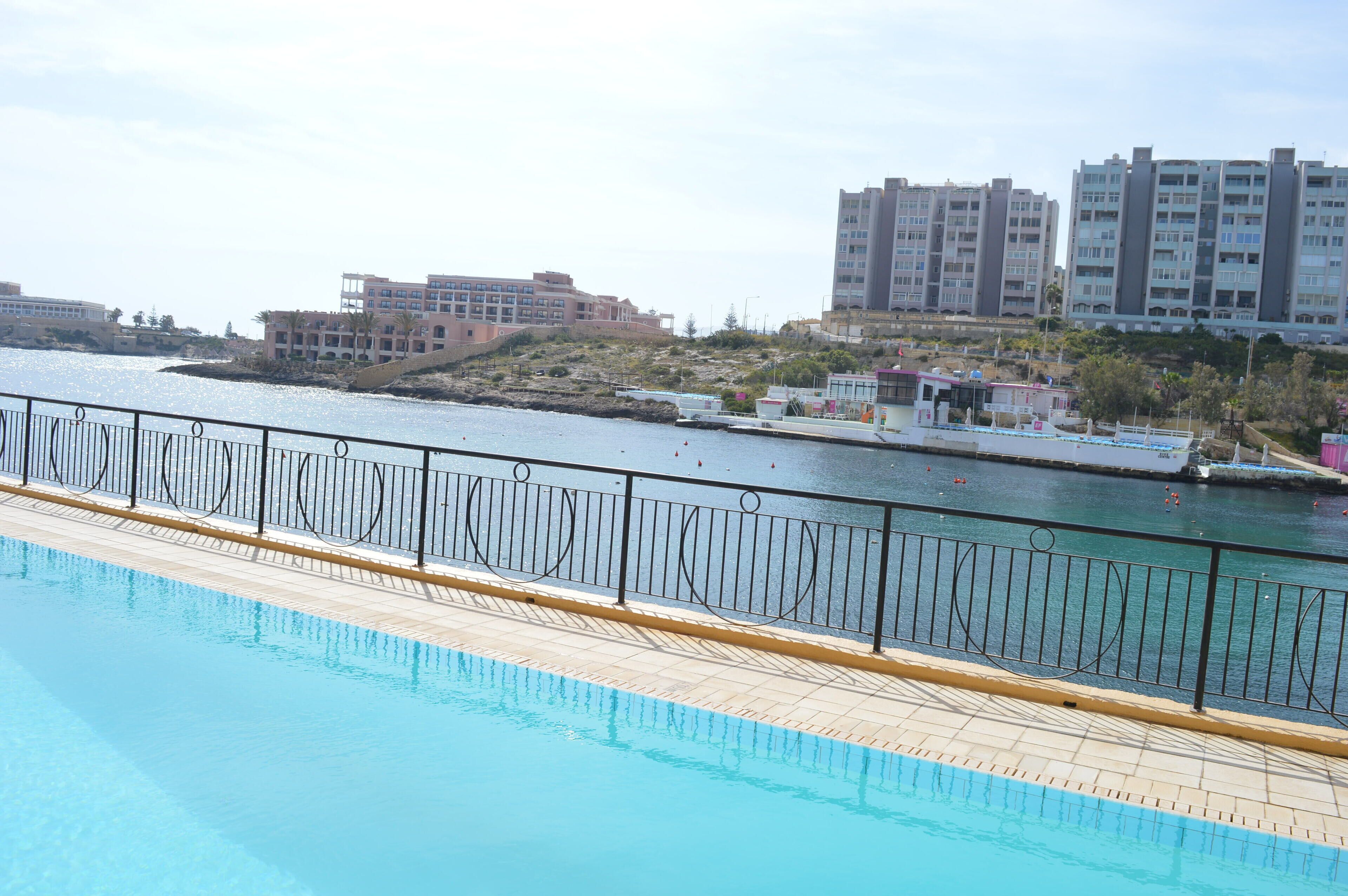 A swimming pool with a metal railing, a waterfront view, and buildings in the background.