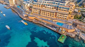 Vista aérea de un hotel costero con varias edificaciones, un puerto deportivo con barcos y una piscina.